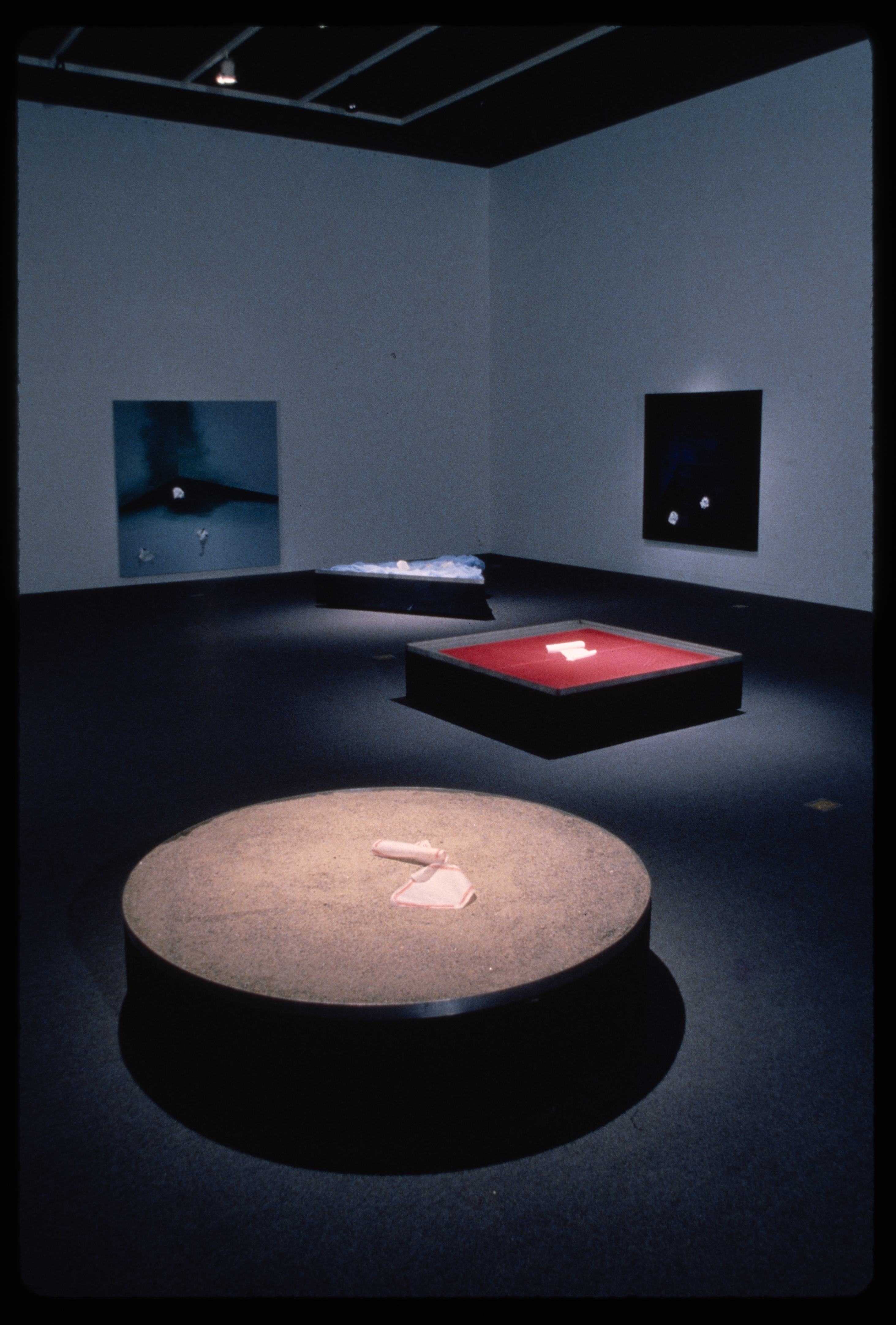 Circular, square, and triangular pedestals are spotlit in the gallery. Blue toned works hang behind them on the walls.