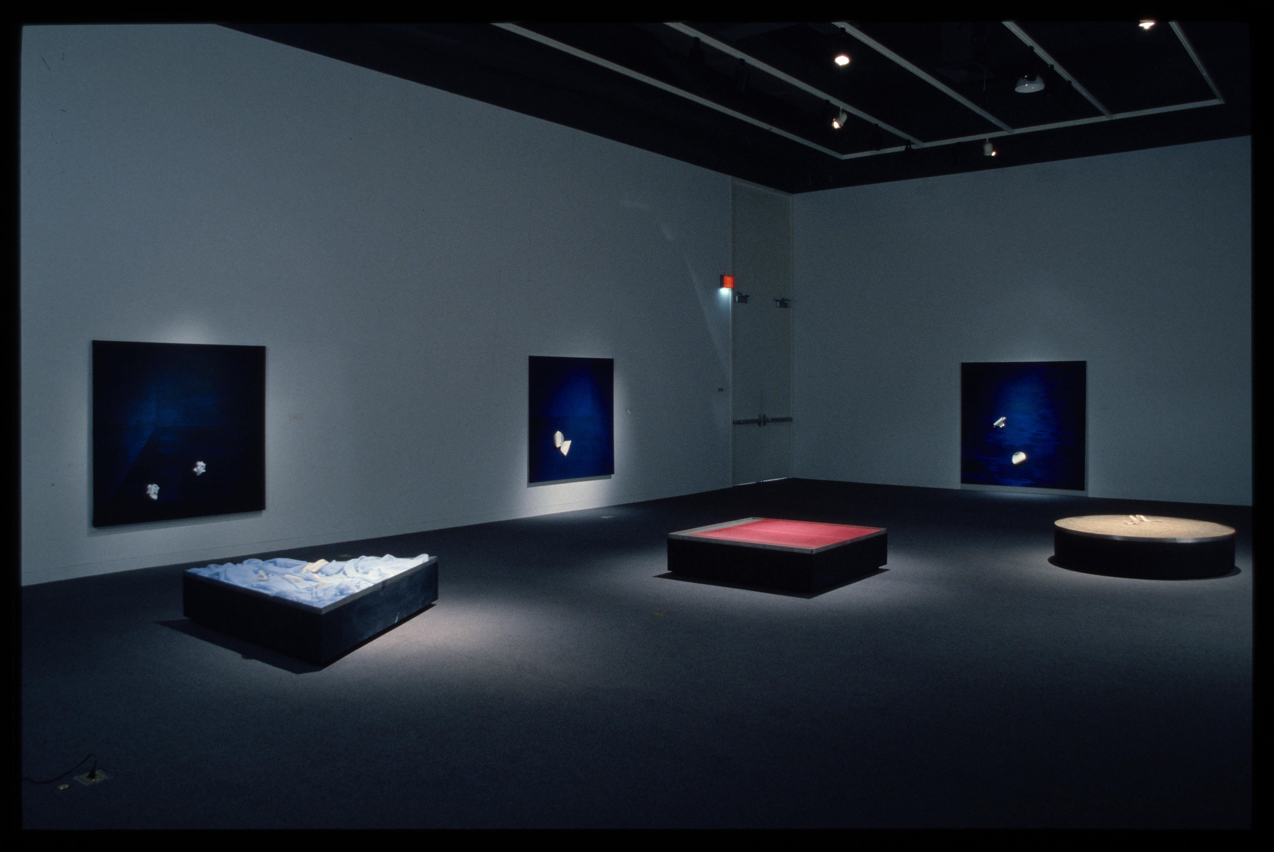 Circular, square, and triangular pedestals are spotlit in the gallery. Blue toned works hang behind them on the walls.