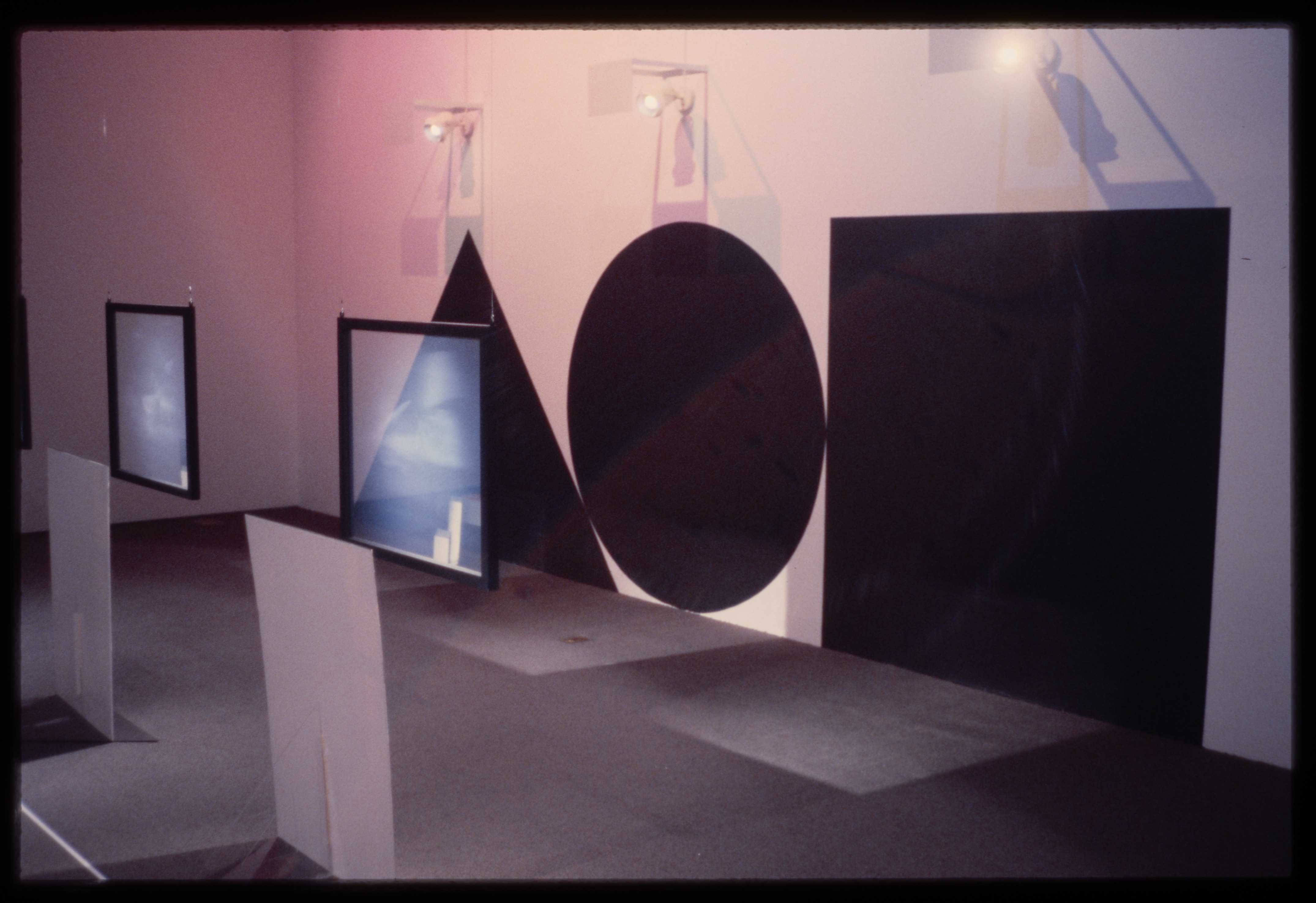 Projection screens hang before a black triangle, circle and square adhered to the wall. Pink lights glow in the background.