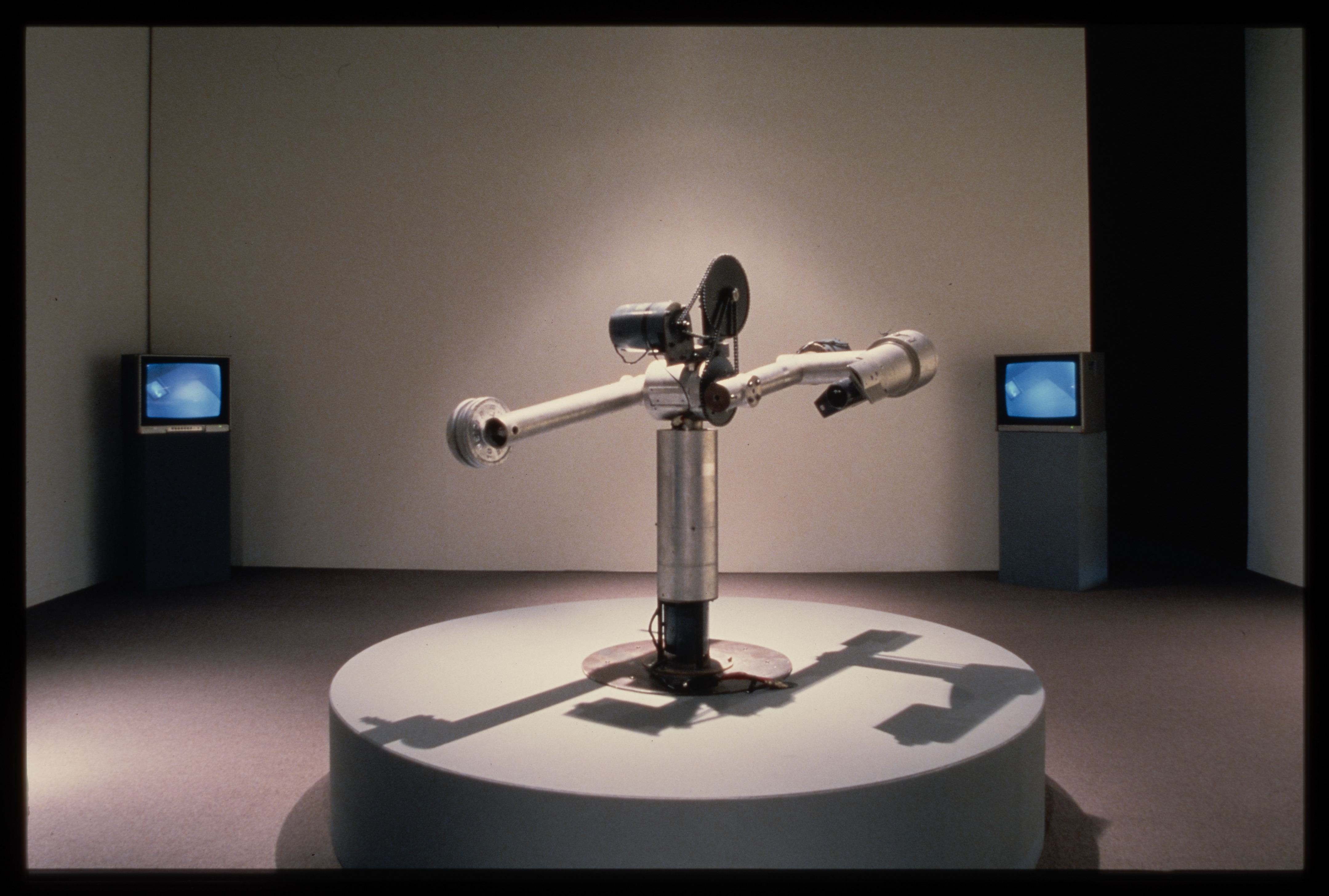 A mechanism sits atop a circular pedestal. A silver cylinder acts as a base while two cylindrical arms are attached by gears.