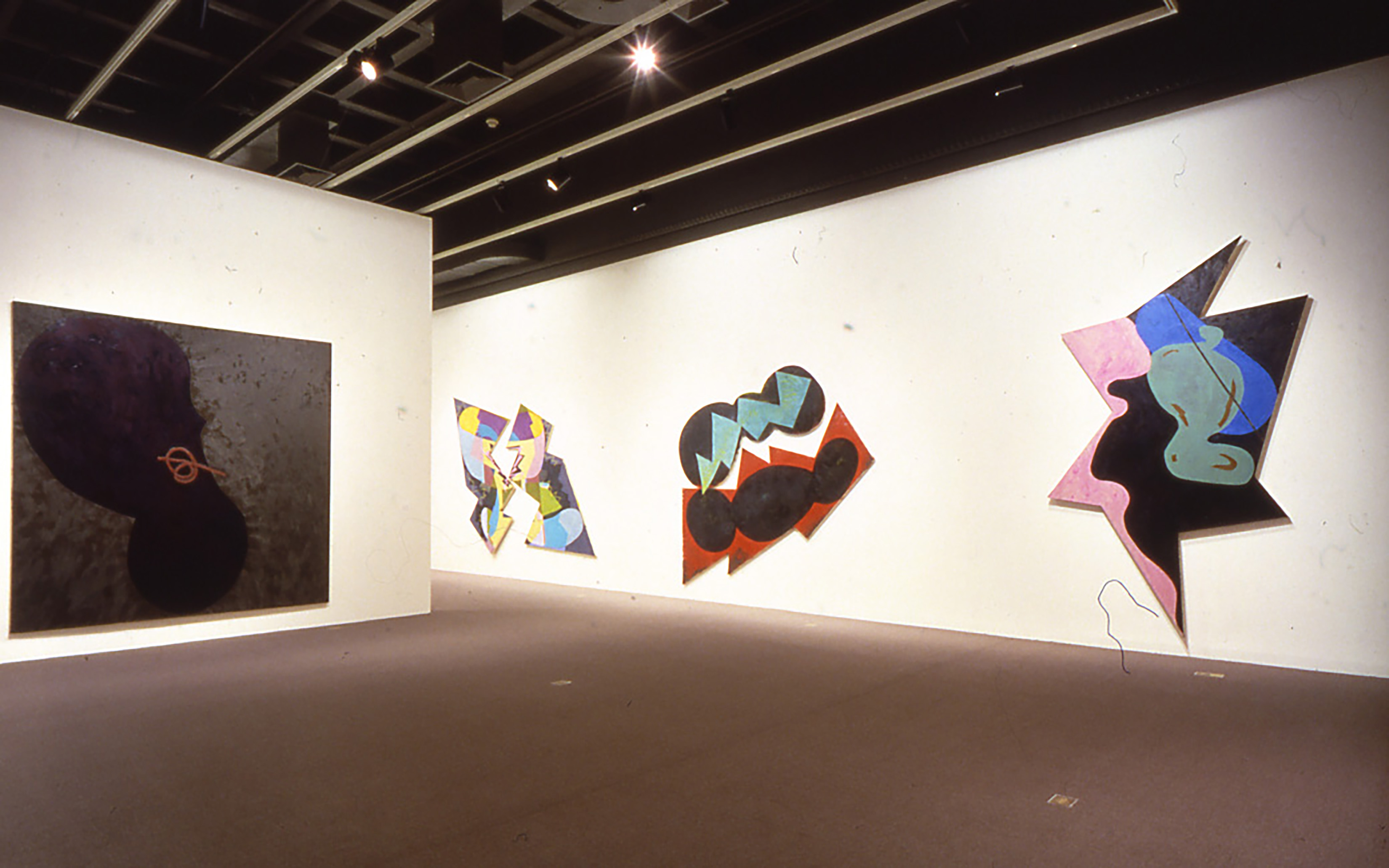 A large dark rectangle shaped painting is hung on one wall and 3 vivid abstract multi shaped paintings are hung on another.