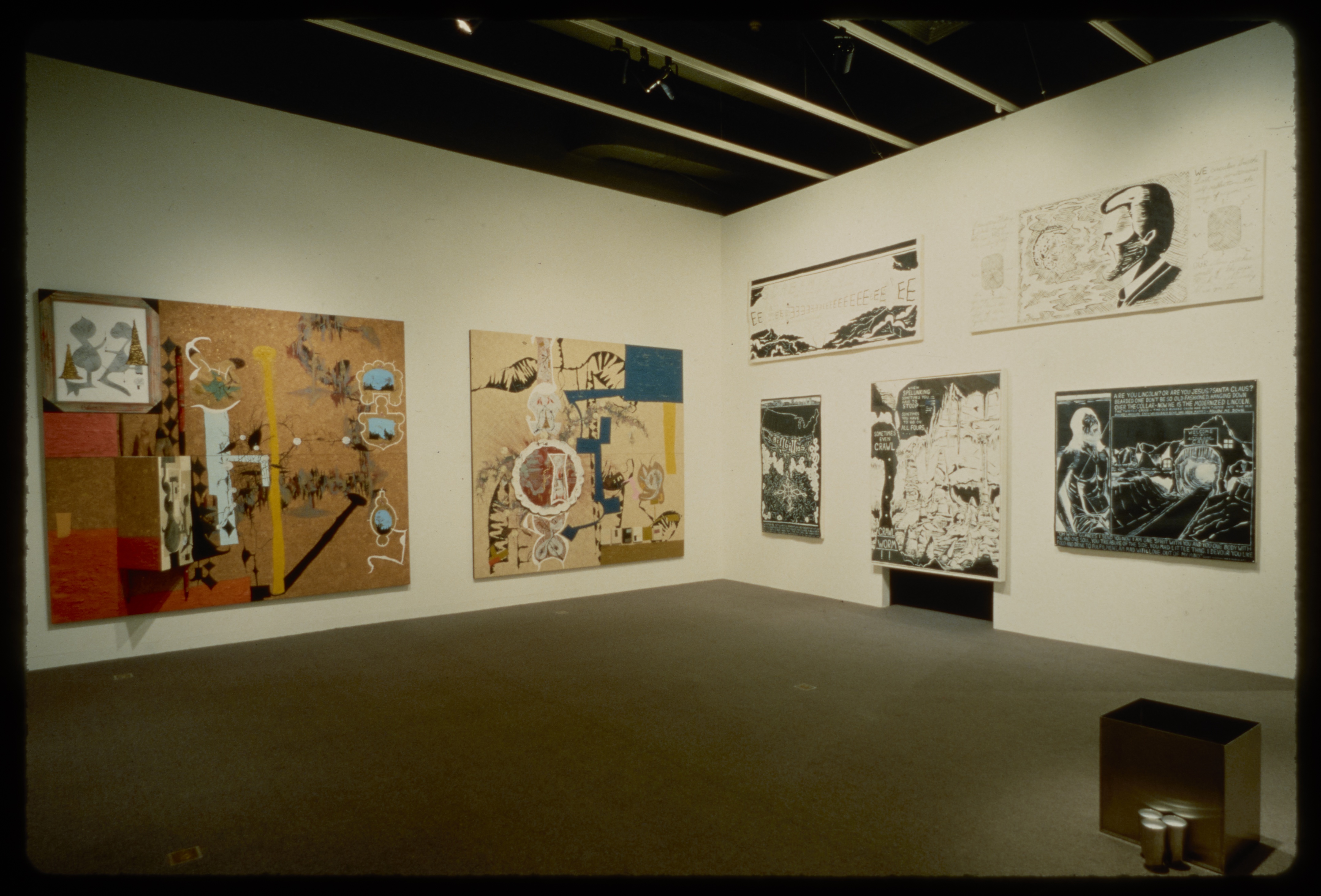 Two large wooden canvases with abstract colorful configurations are hung adjacent to five black and white comic style paintings.