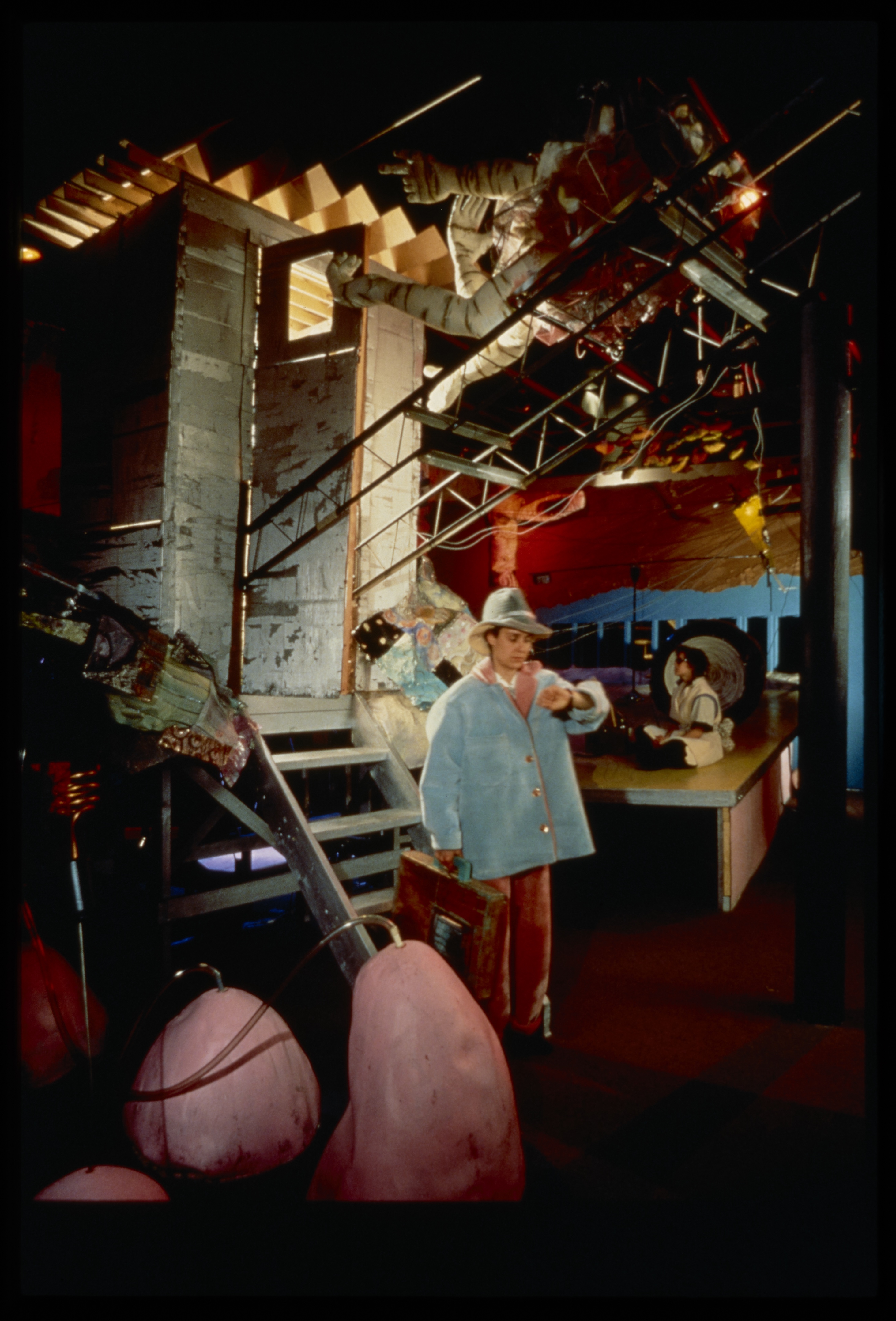 Person in hat and blue coat look at watch while woman sits on a platform in an abstract mechanical landscape installation.