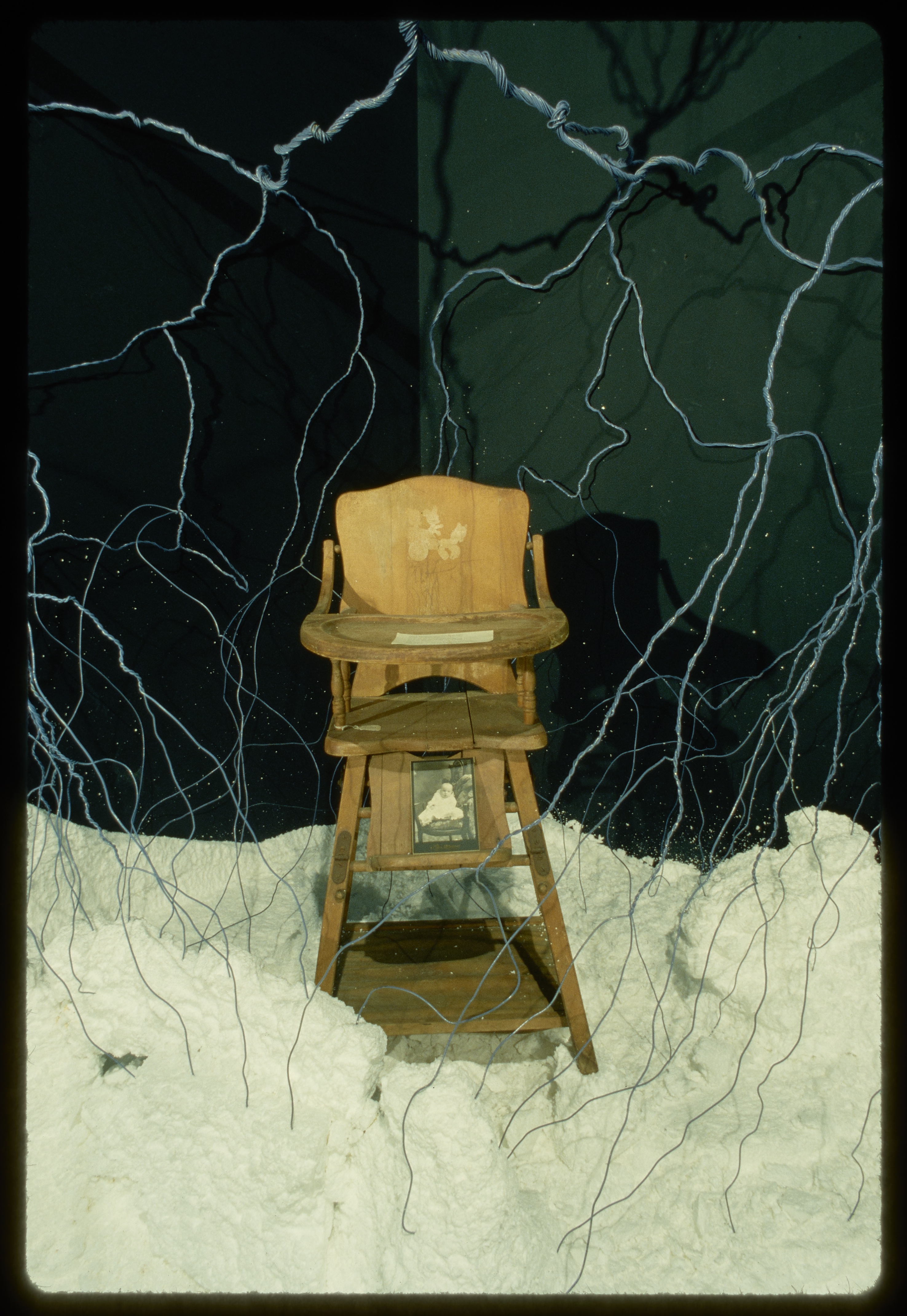 A wooden highchair sits in white material made to look like snow. On the chair is an old black and white image of a child.