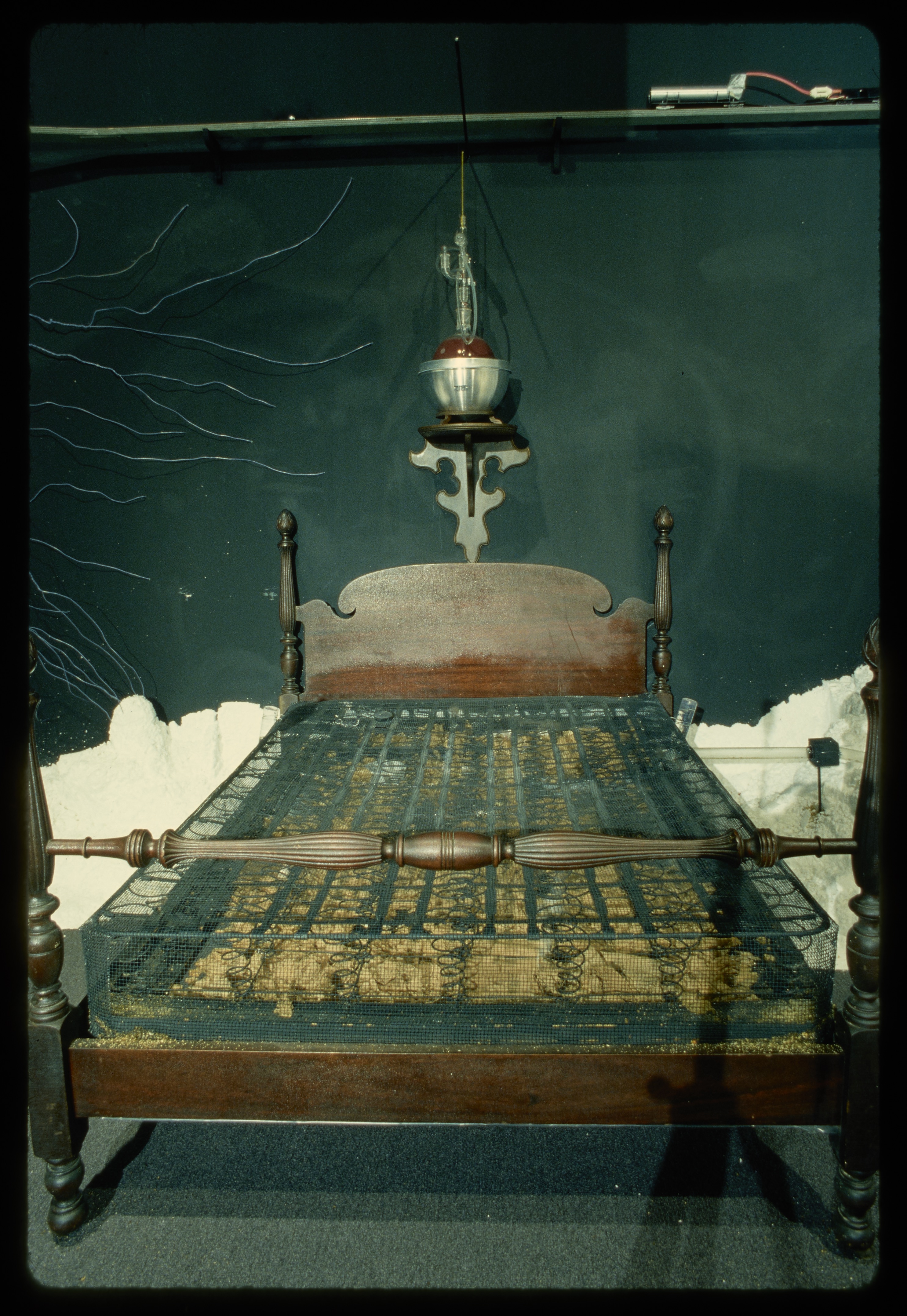 A bed with just the box spring sits against the wall. Above the bed is a mounted sculpture with a red orb in the center.