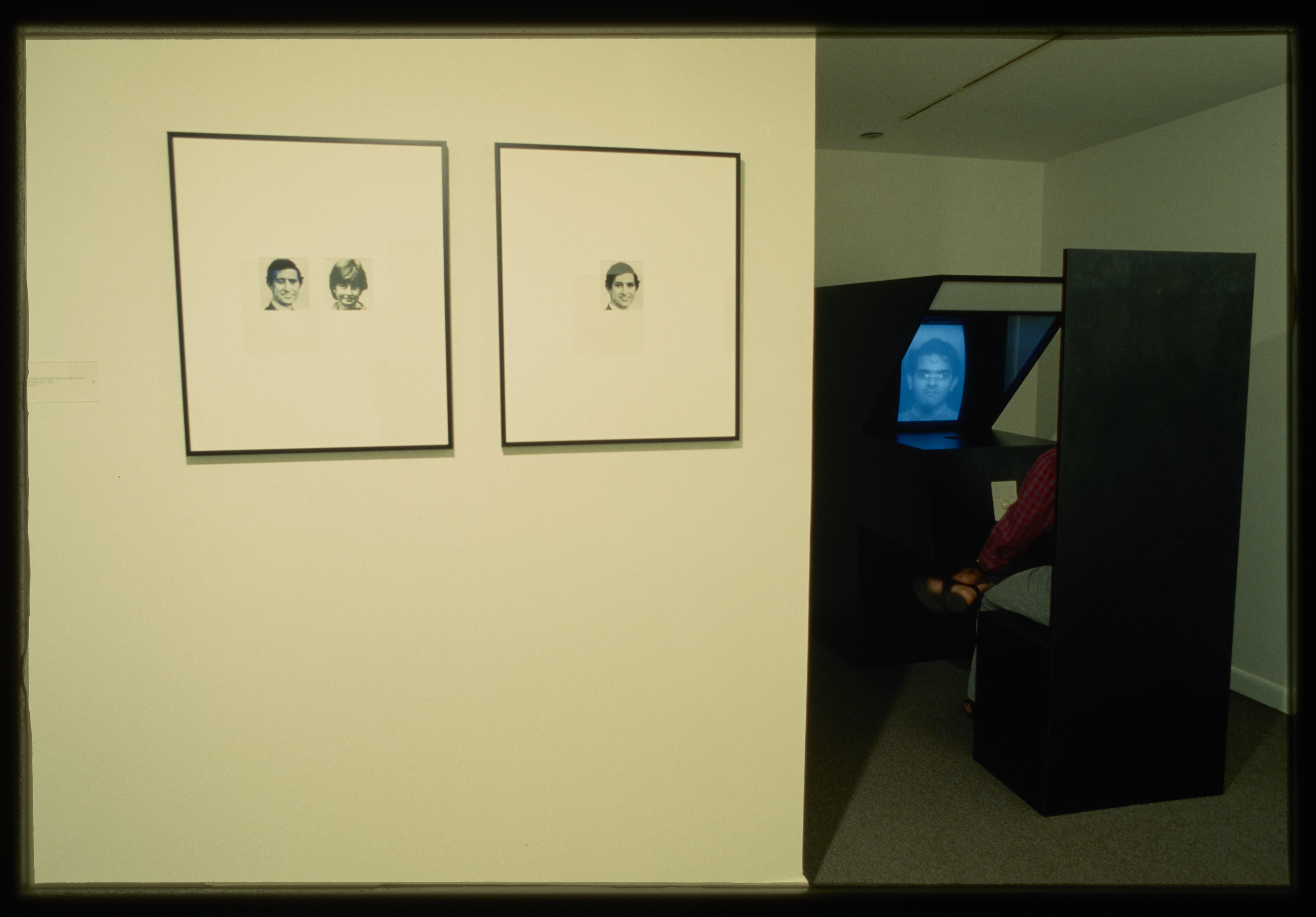 Two images hang on the gallery wall while a photobooth is set up in a dark corner of the gallery.