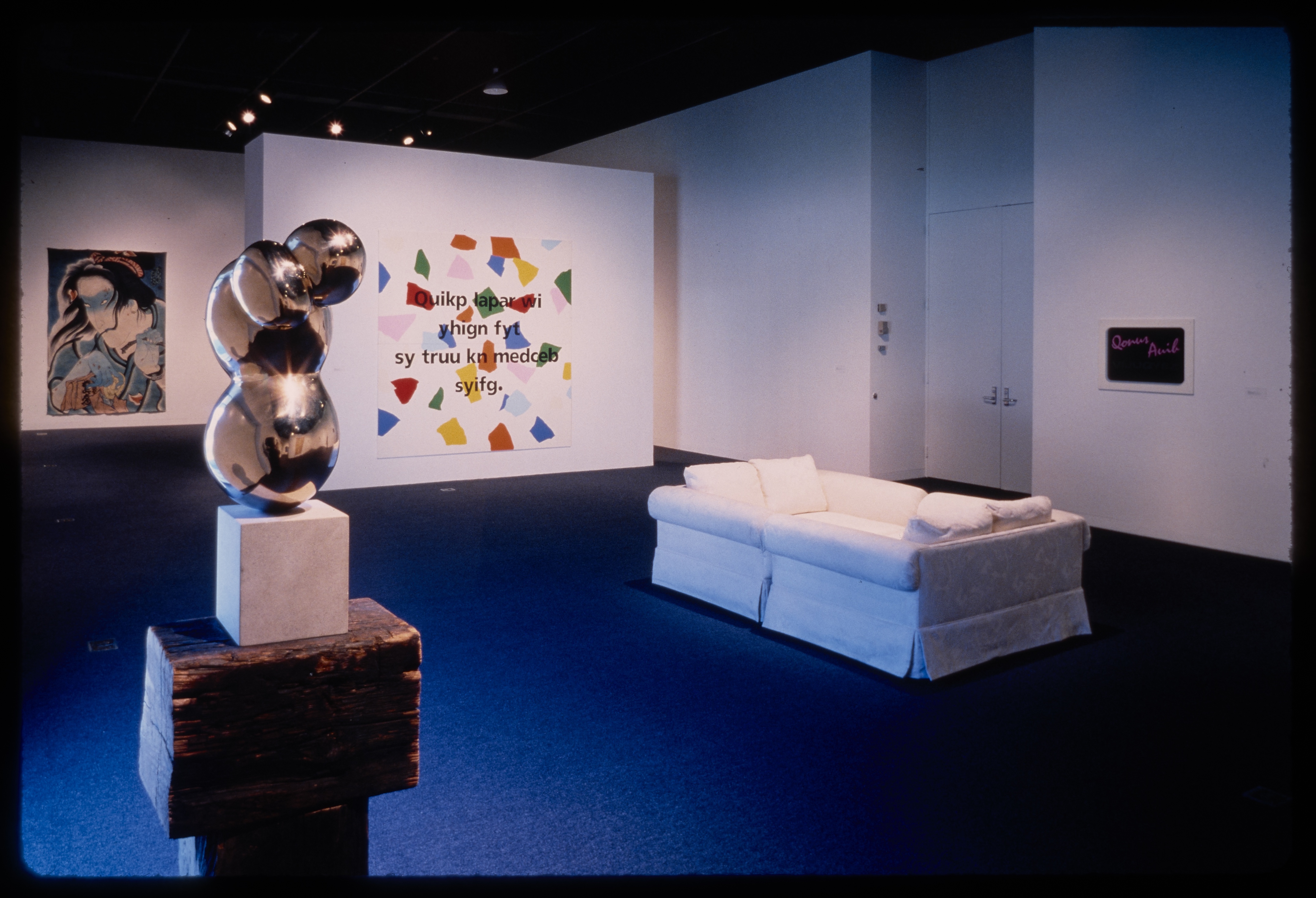 An abstract sculpture stands in the foreground while a white couch is settled behind. Hanging works are on the surrounding walls.