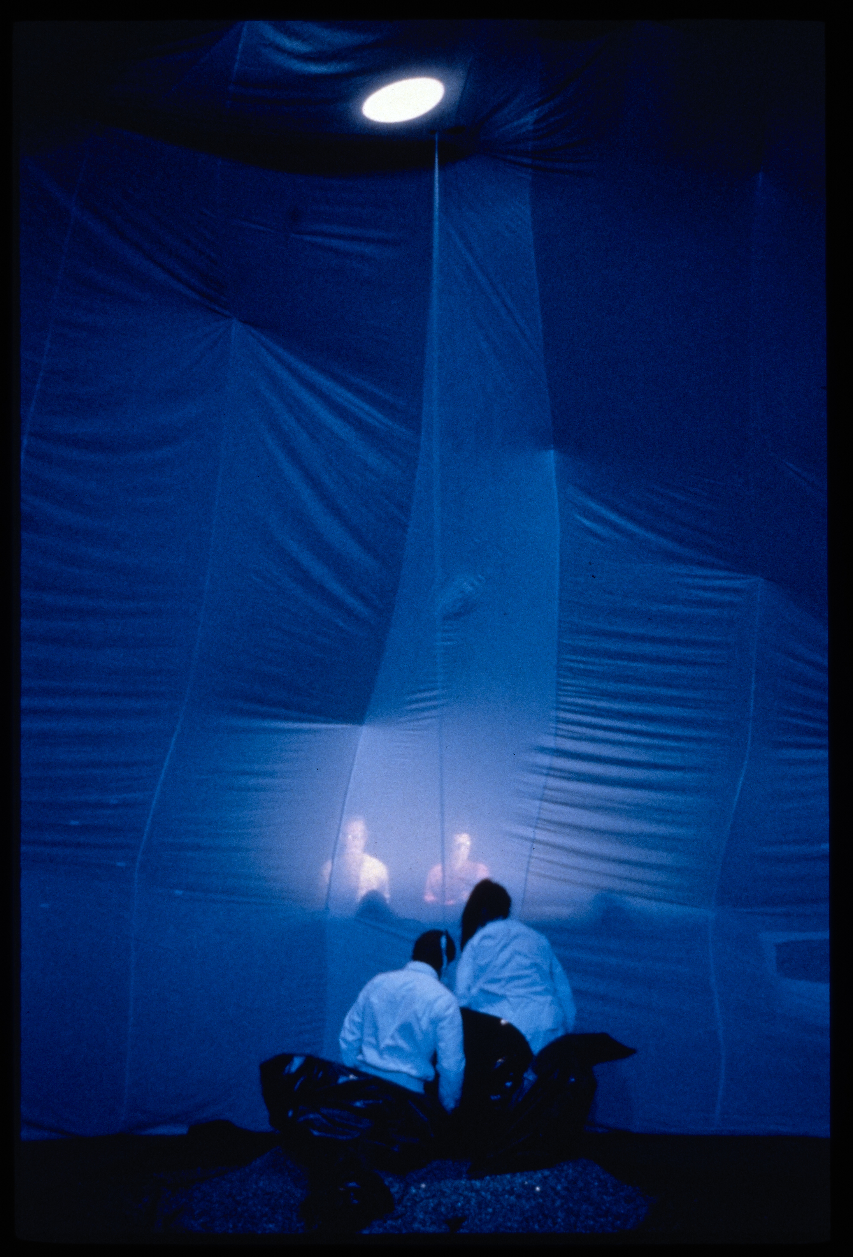 Two performers dressed in white emerge from encasings while watching a projection of two people before them.