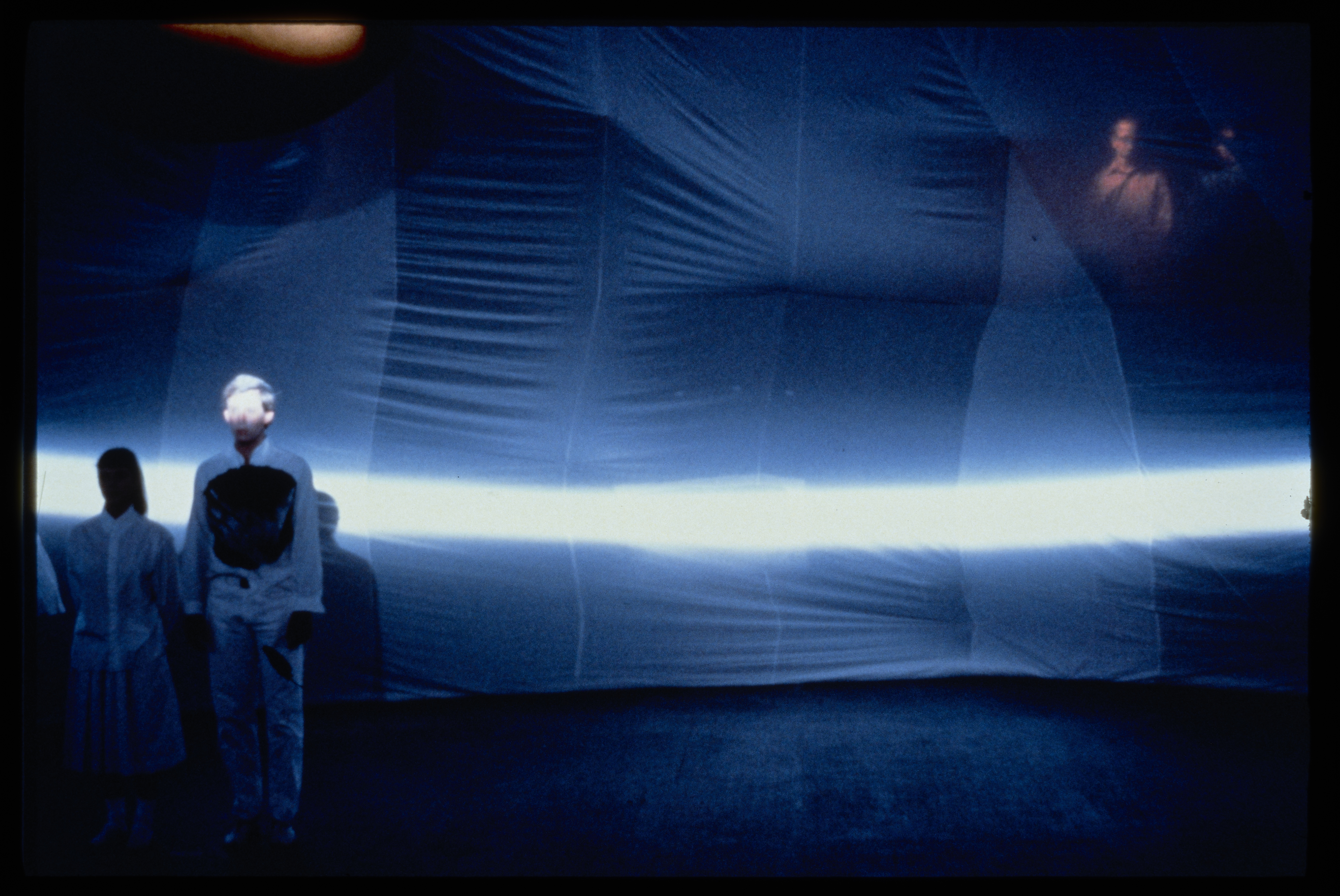 Two performers stand to the left of the image. Sheets hung behind them are illuminated by a blue streak of light.