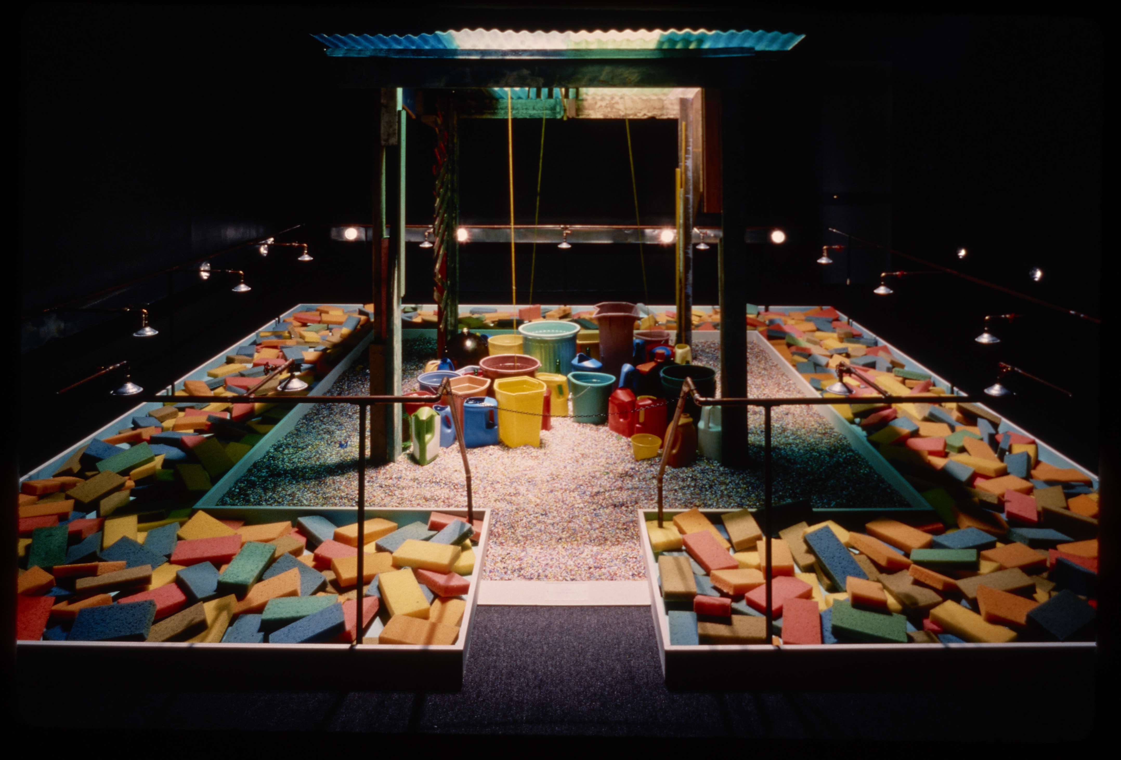 Wooden playset with spotlight on plastic sand castle buckets in sand, surrounded by troughs filled with colorful wooden bricks.
