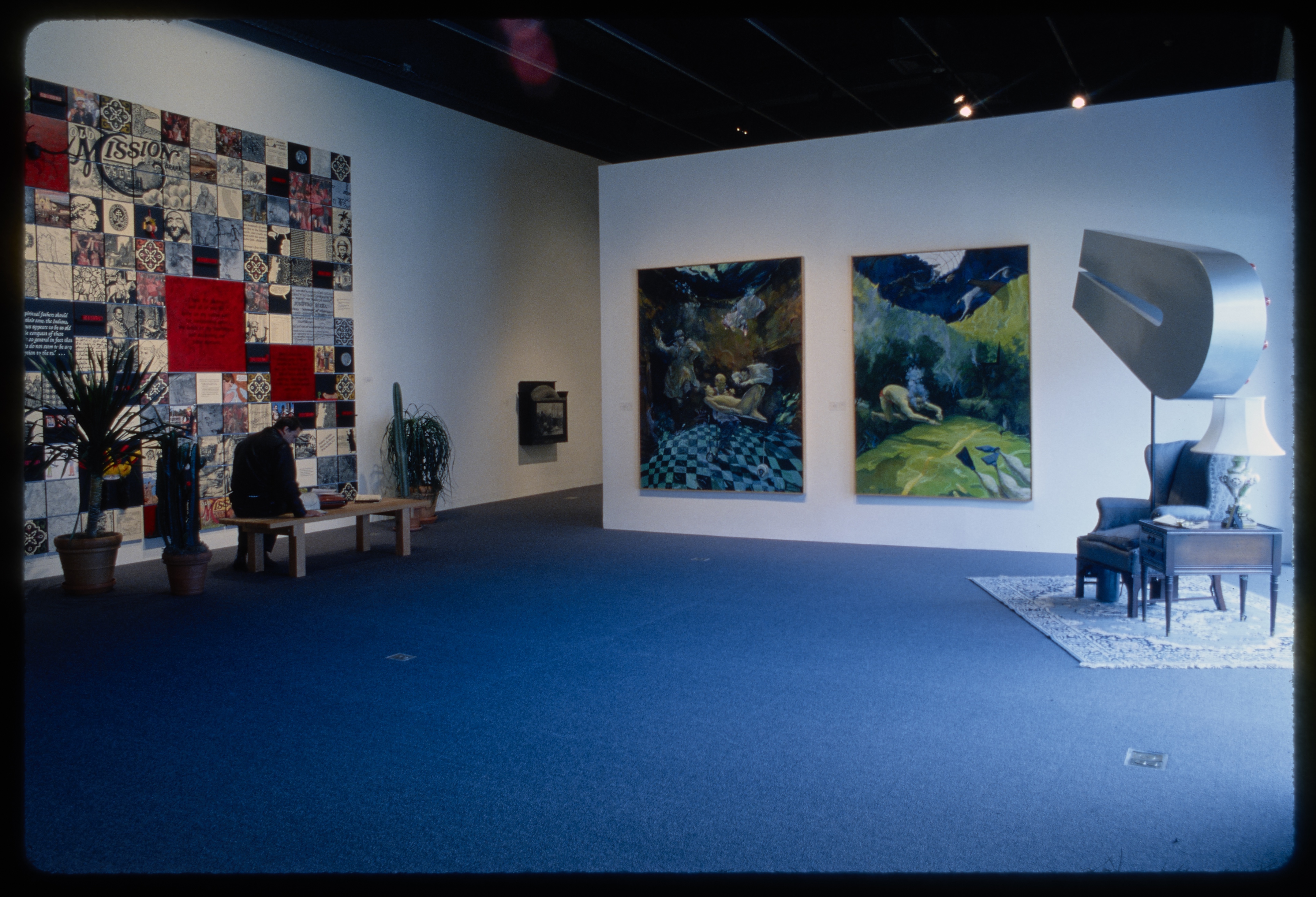 Large patchwork tapestry hangs on wall adjacent to two large natural paintings. Magnet above armchair sits in corner of room.