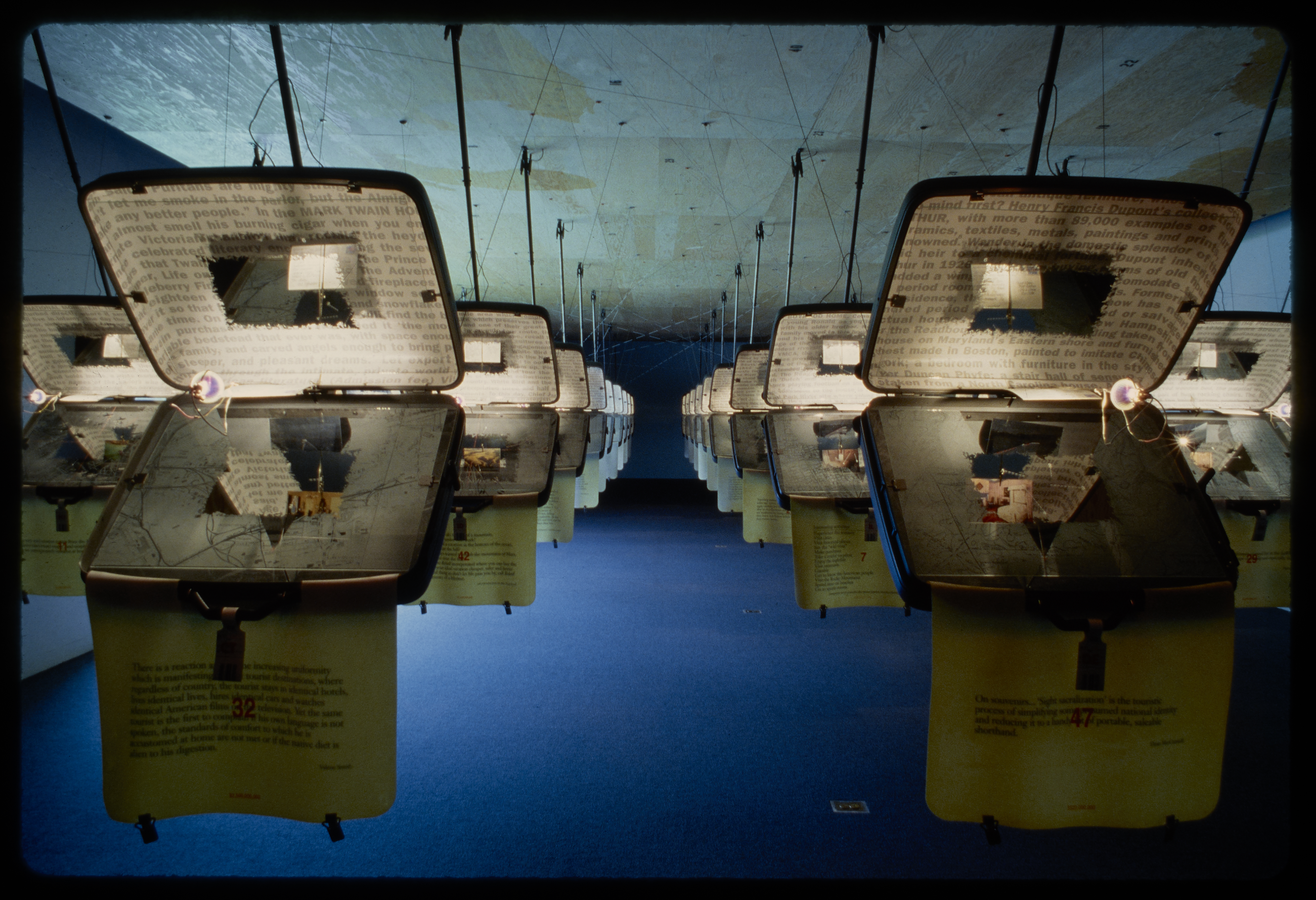 Identical opened suitcases are hung from the ceiling in rows. Each has text projected on the inside.