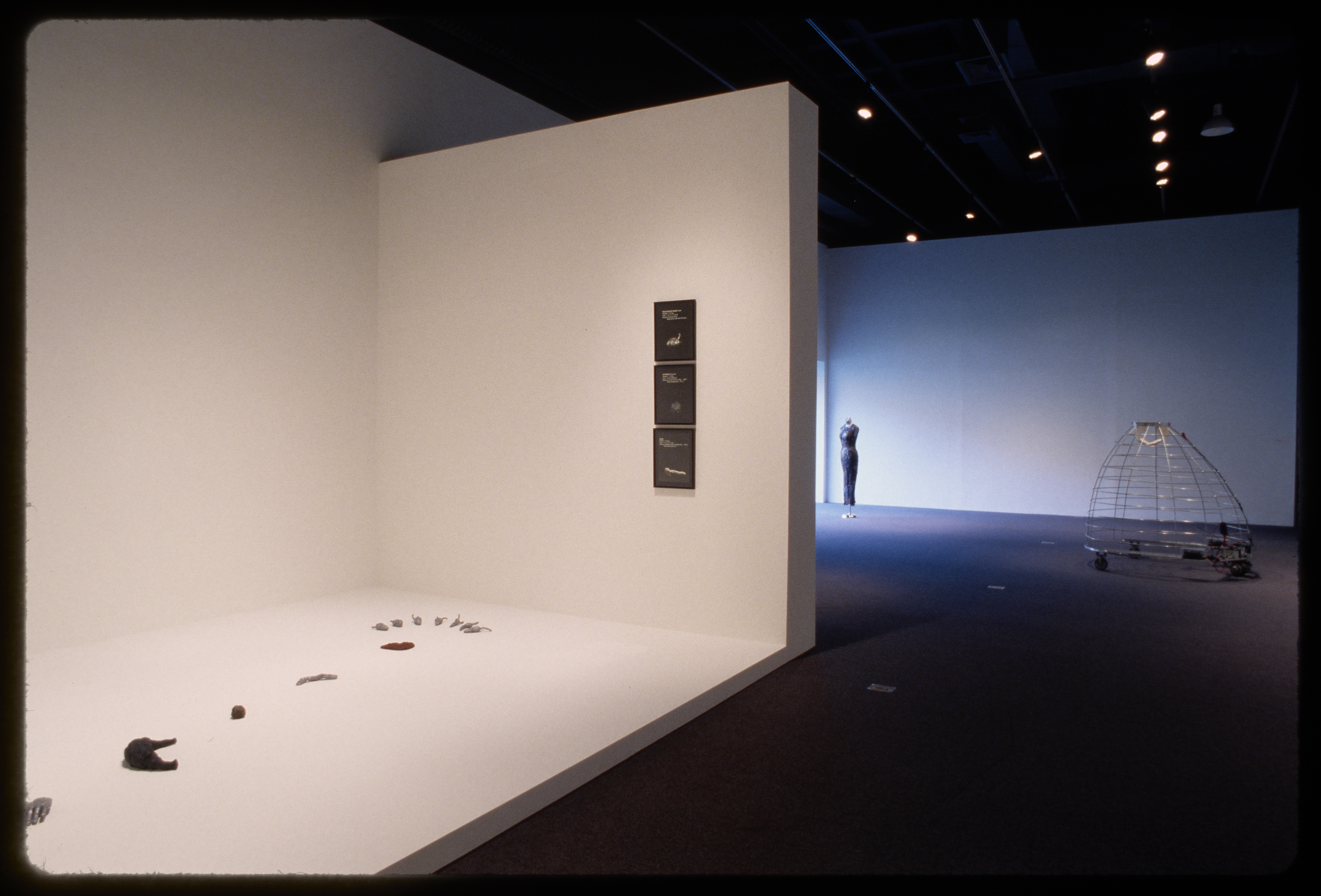 A raised floor holds small objects at the entrance of the gallery. A wire skirt and a mannequin stand further in the gallery.