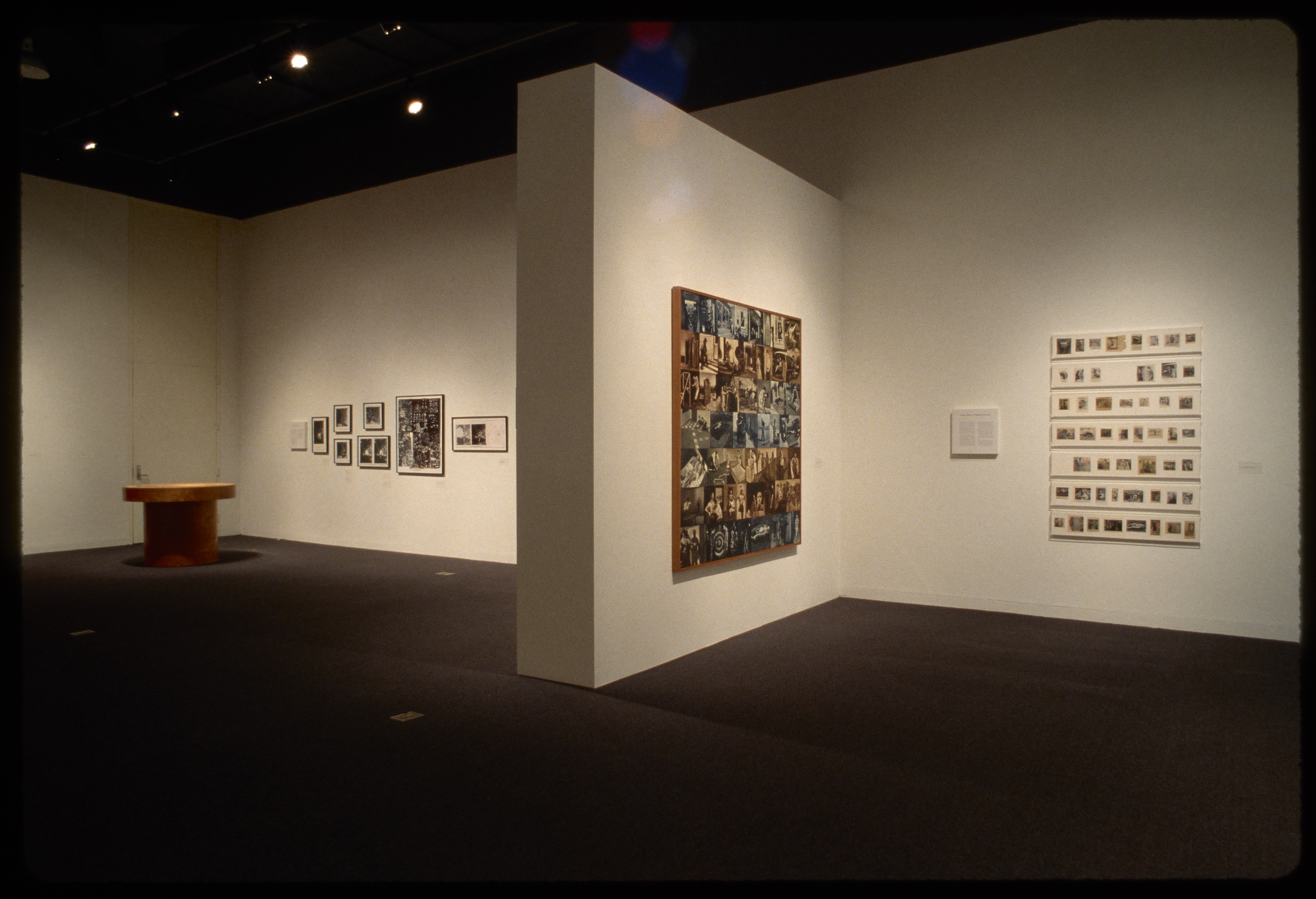 Wall separates art spaces: one with patchwork sepia photos and fifty photos, other with eight framed photos and wooden table.