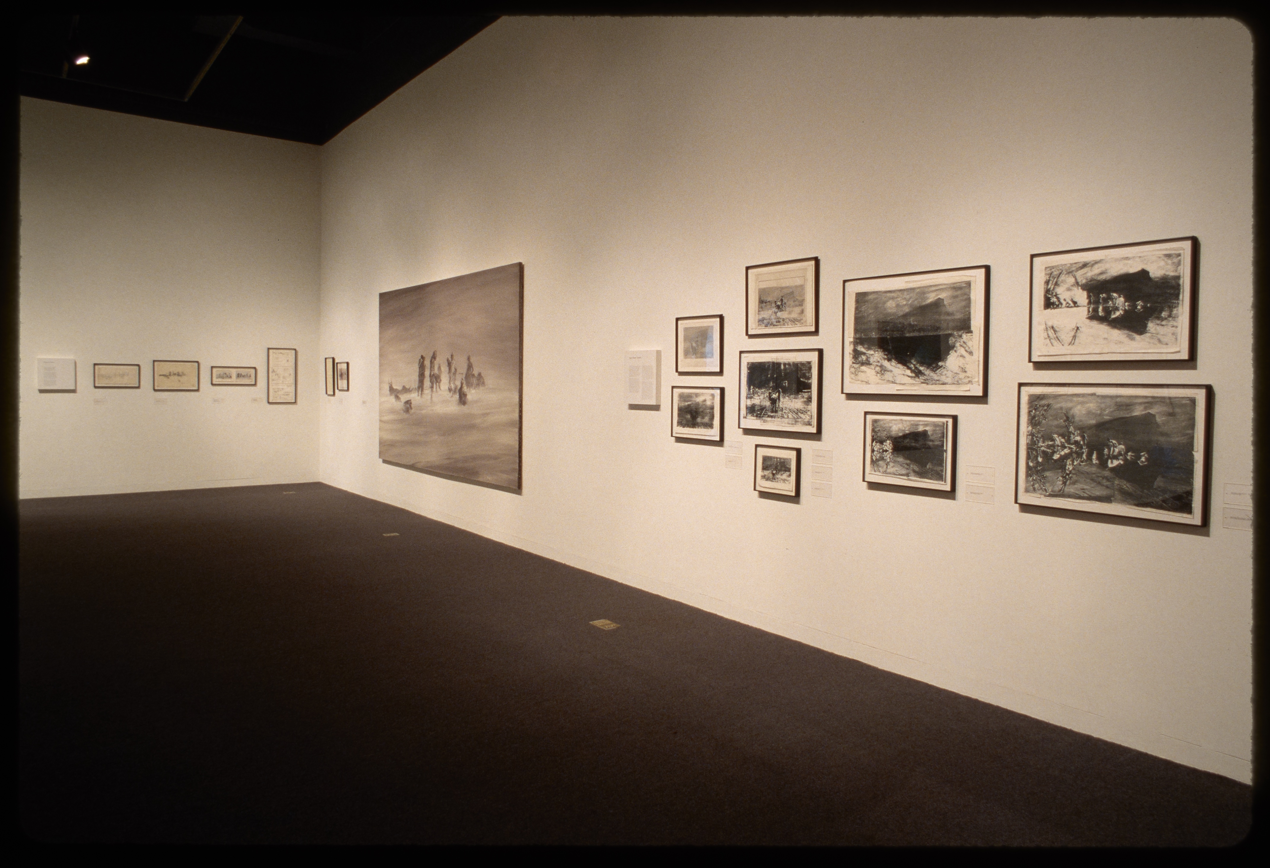 Framed text hangs adjacent to large monochrome oil painting of nomads in a desert and nine framed black and white photographs.