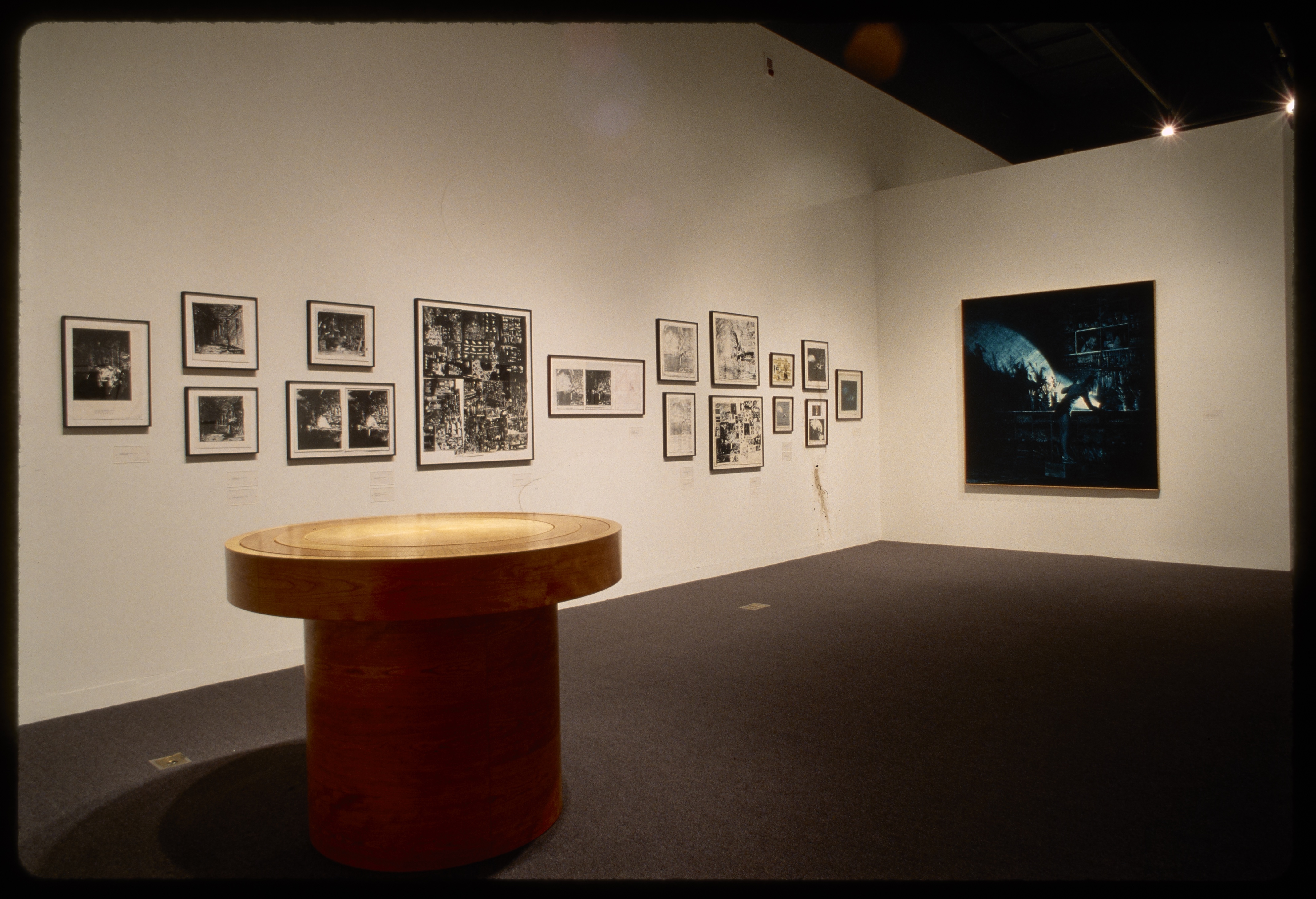 Circular wooden table sits gallery with sixteen framed black and white photographs and large square photograph on walls.