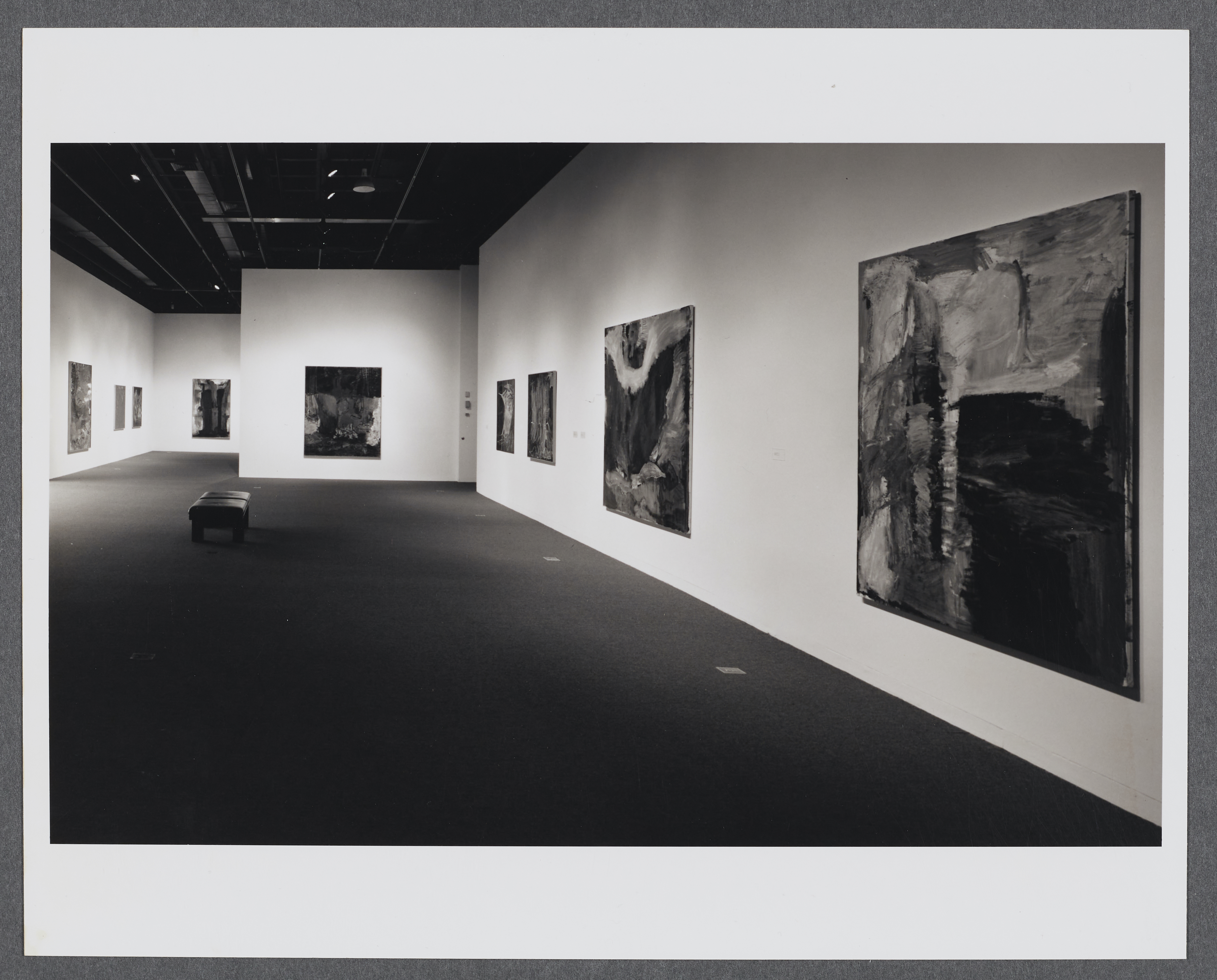 Large abstract paintings line the gallery walls as a bench sits in the center of the gallery.   