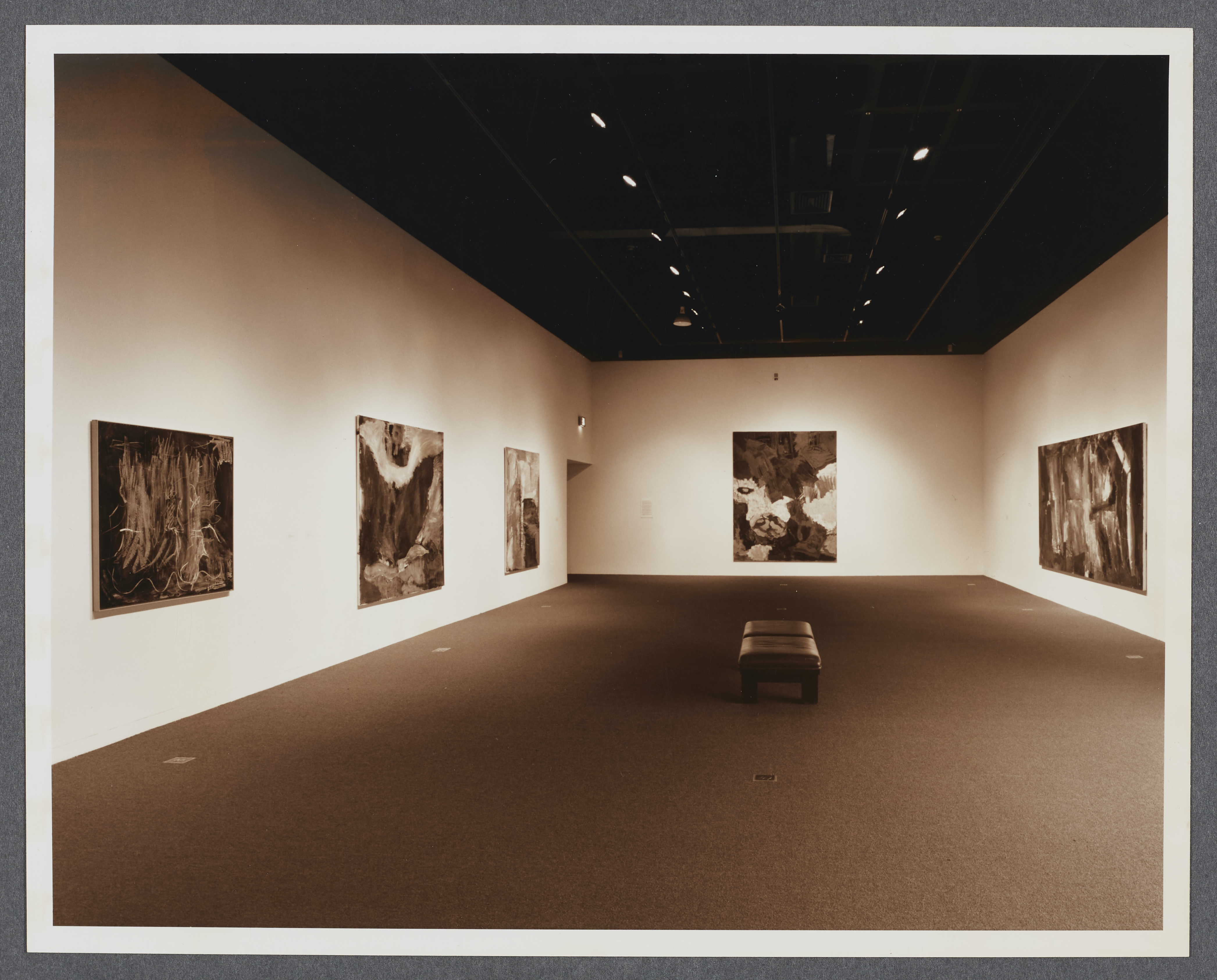 Large abstract paintings line the gallery walls as a bench sits in the center of the gallery. 