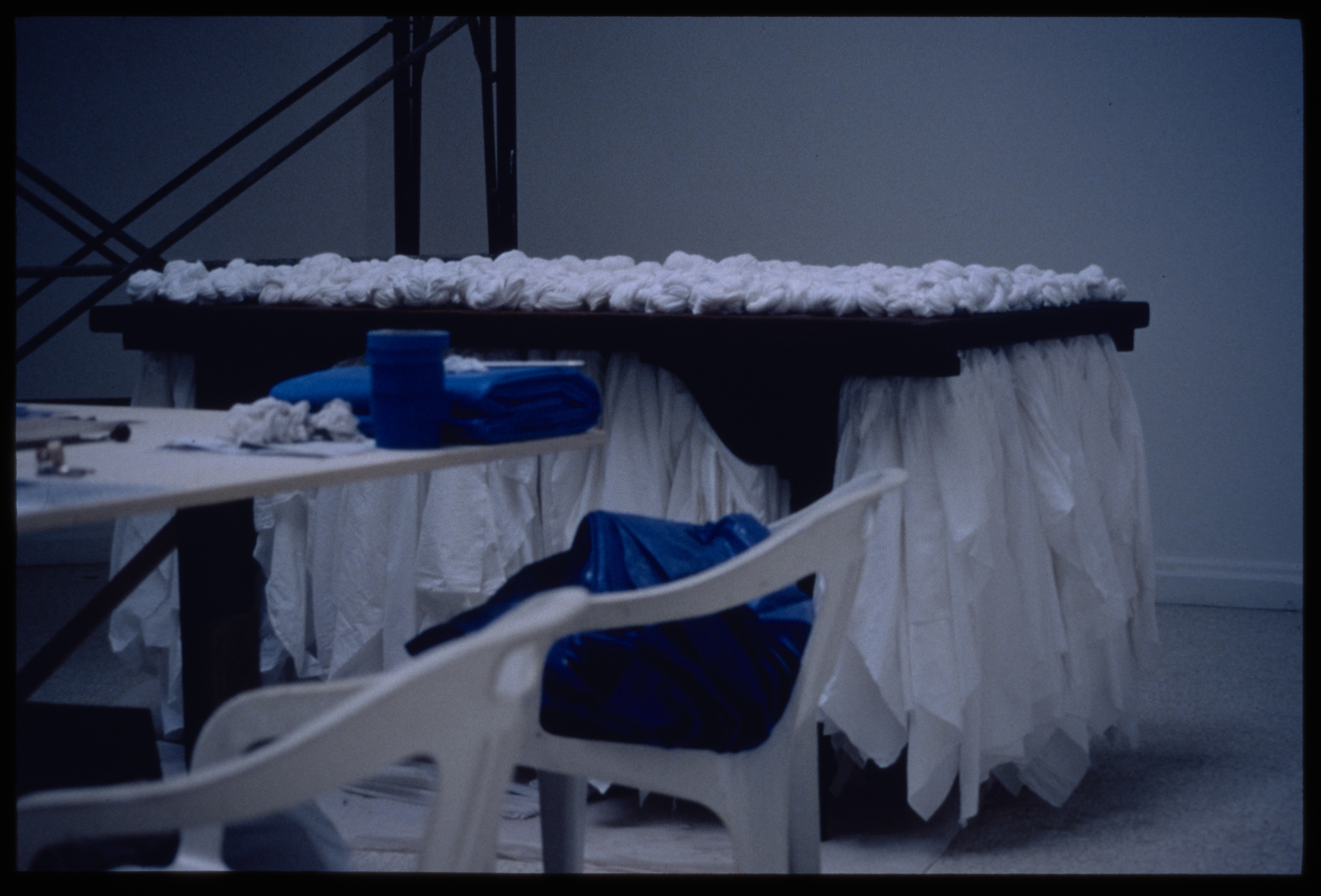 Black table with white bunched fabric on top and bottom, and white table and chairs with blue objects on them in blue-lit room.