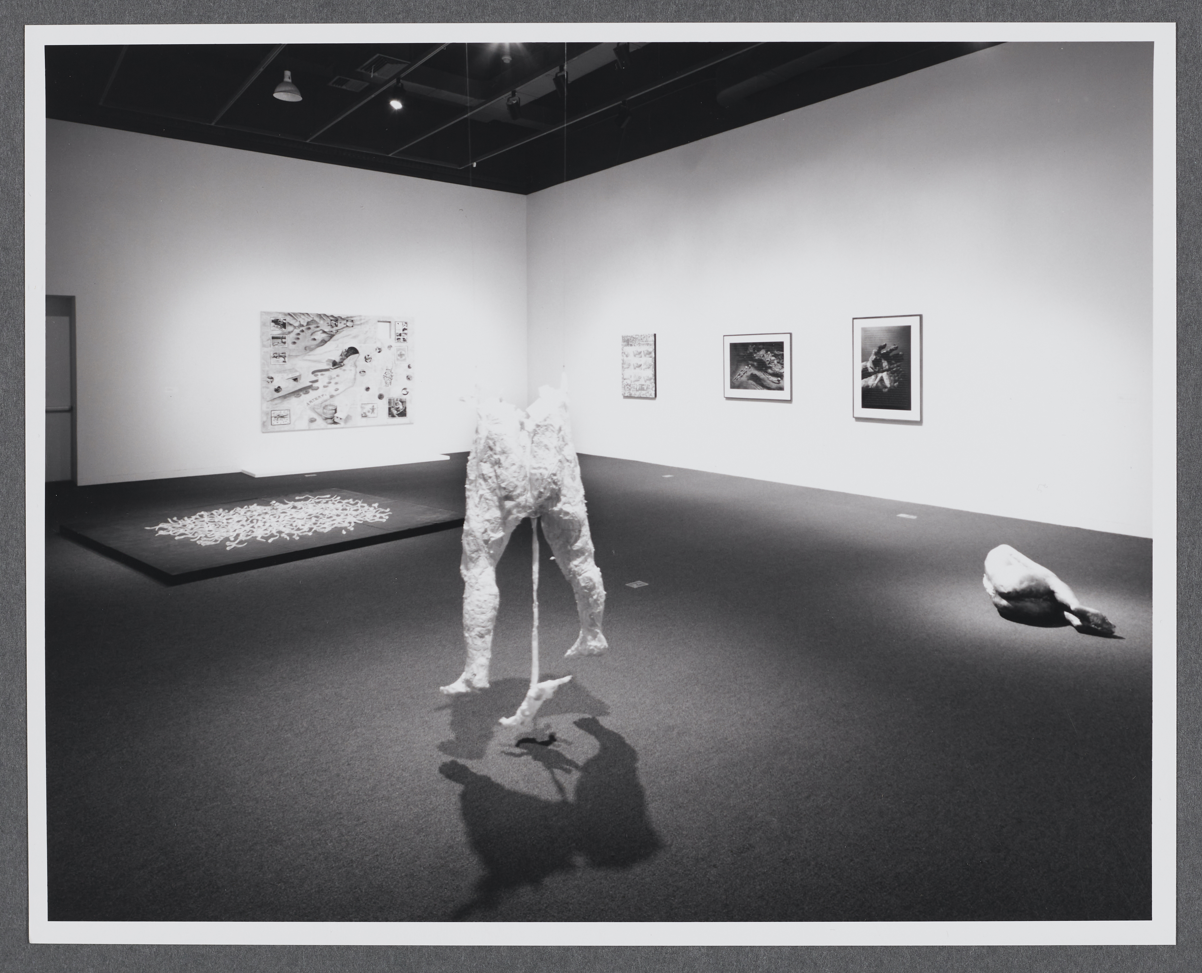 A sculpture of two standing legs is placed in the center of the room. Spotlight from above, the sculpture casts two shadows. 