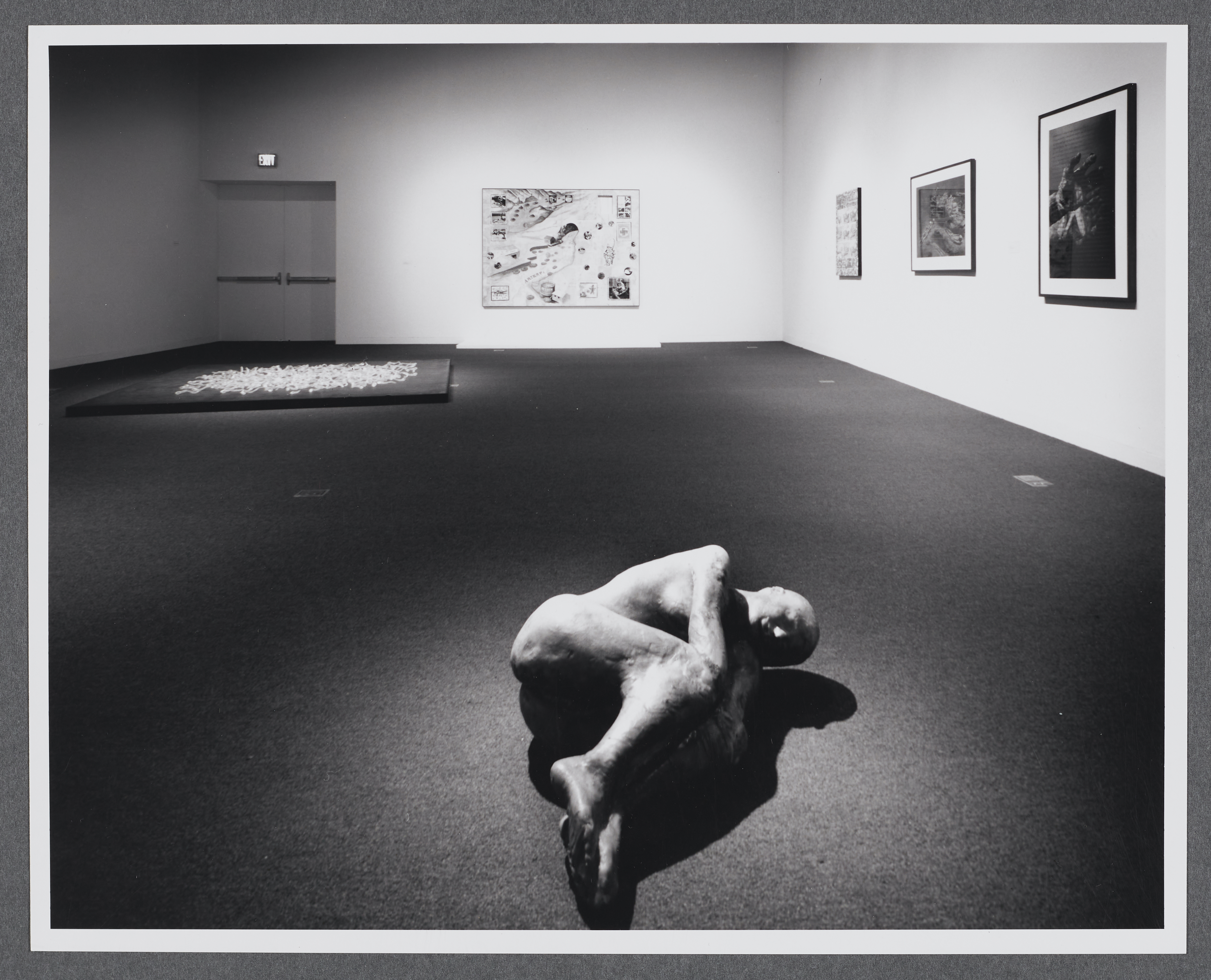 A sculpture of a human figure is curled up on its side. It sits completely alone compared to the other works in the room. 