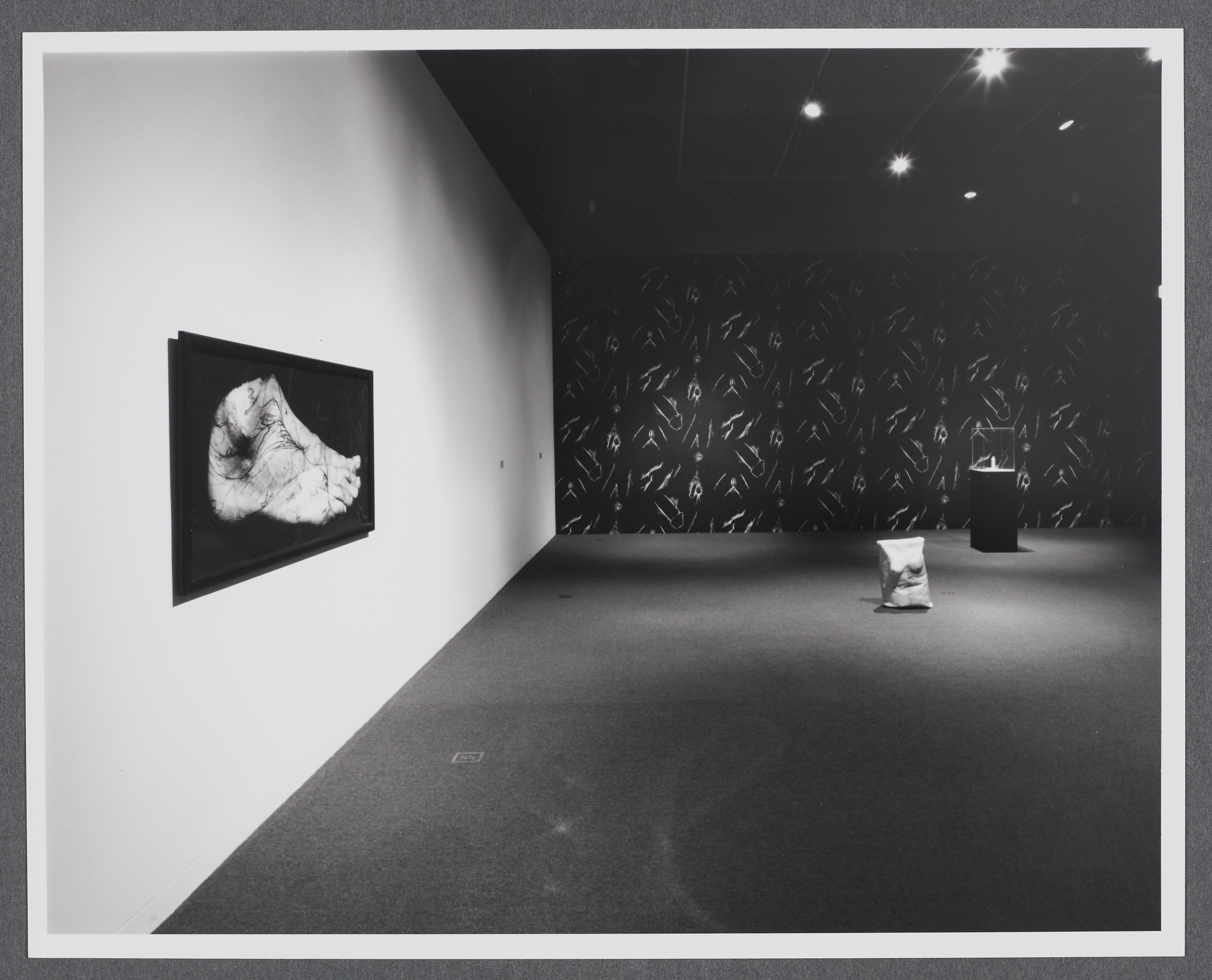 An image of a human foot is hung on the wall. In the distance is a sculpture of a sack made to look like a female torso.  