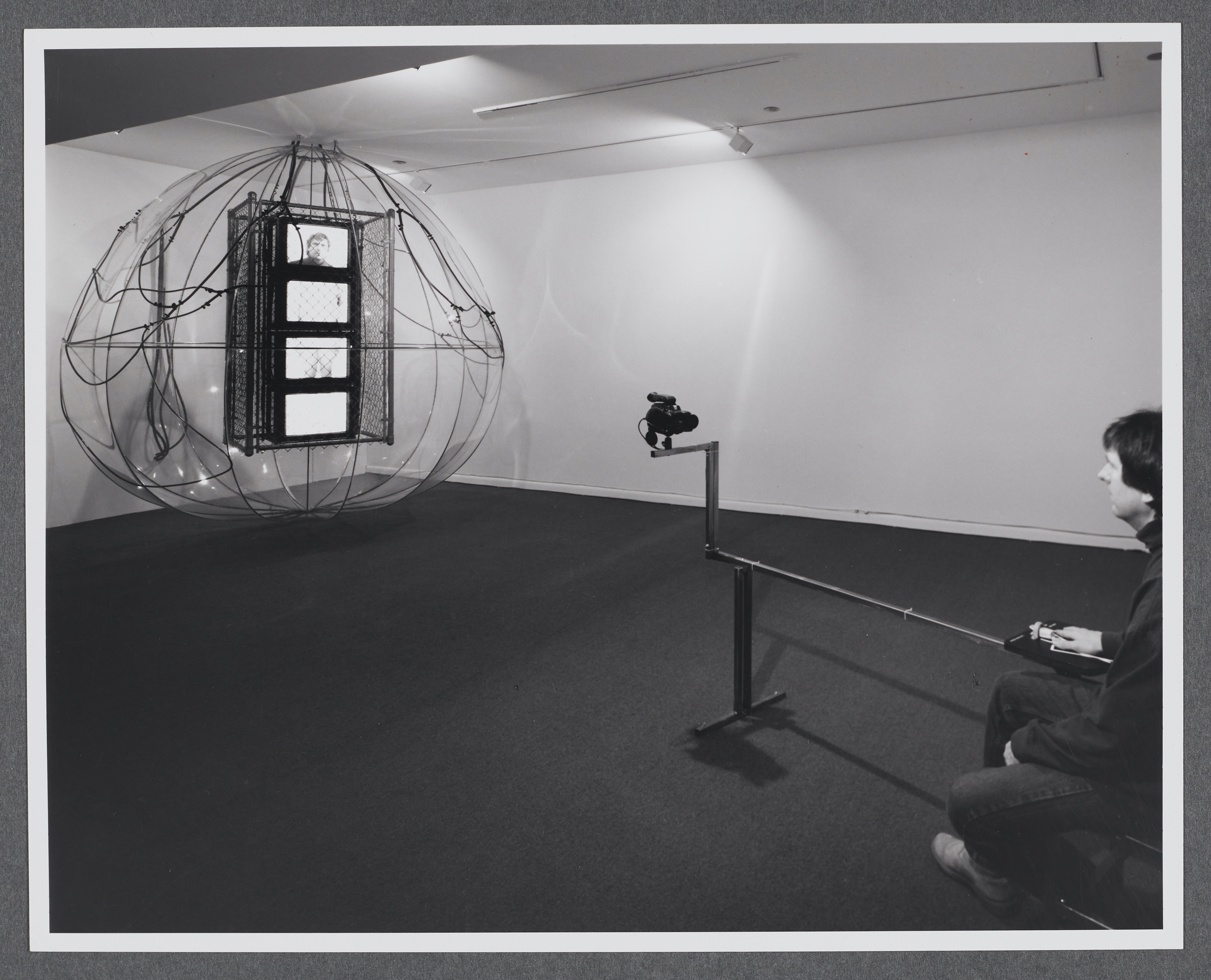 A viewer sits in front of a camera as their image is then projected onto a monitor within the sculpture.