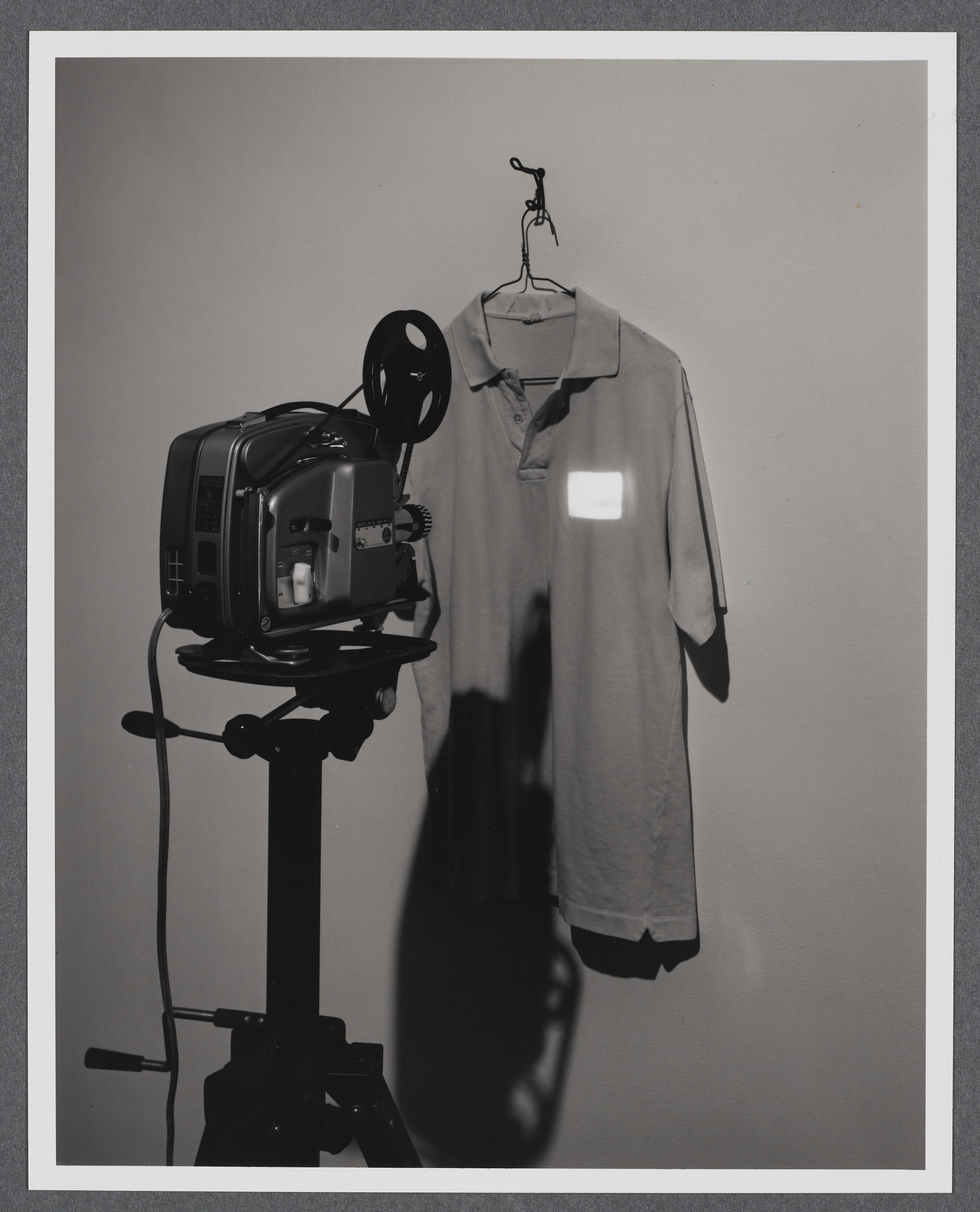 A collared shirt hangs on the gallery wall. A projector projects a tiny image onto the upper right hand side of the shirt.