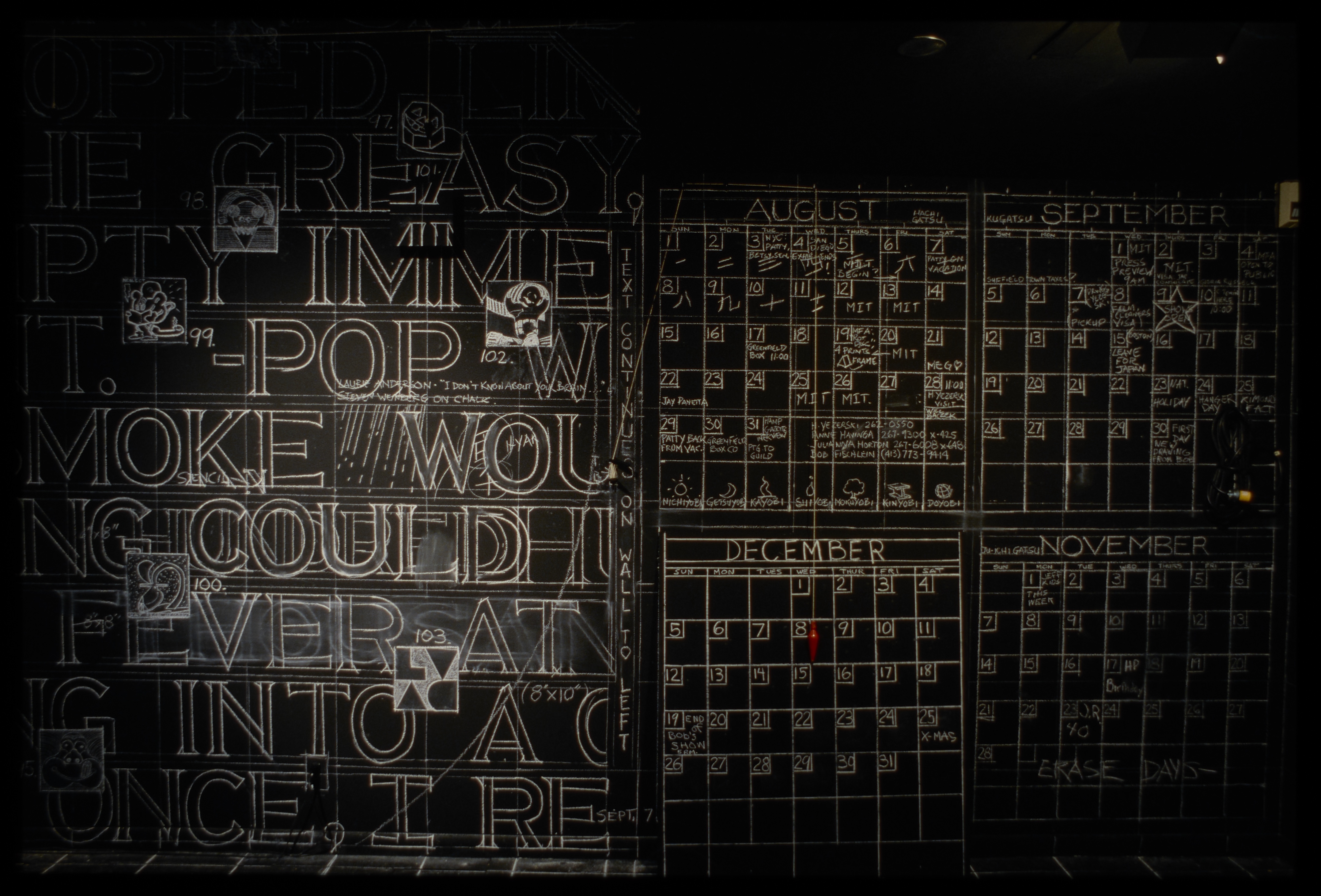 Black wall with spotlit white chalk verbal notations on it: large words on the left, and four notated calendars on the right. 