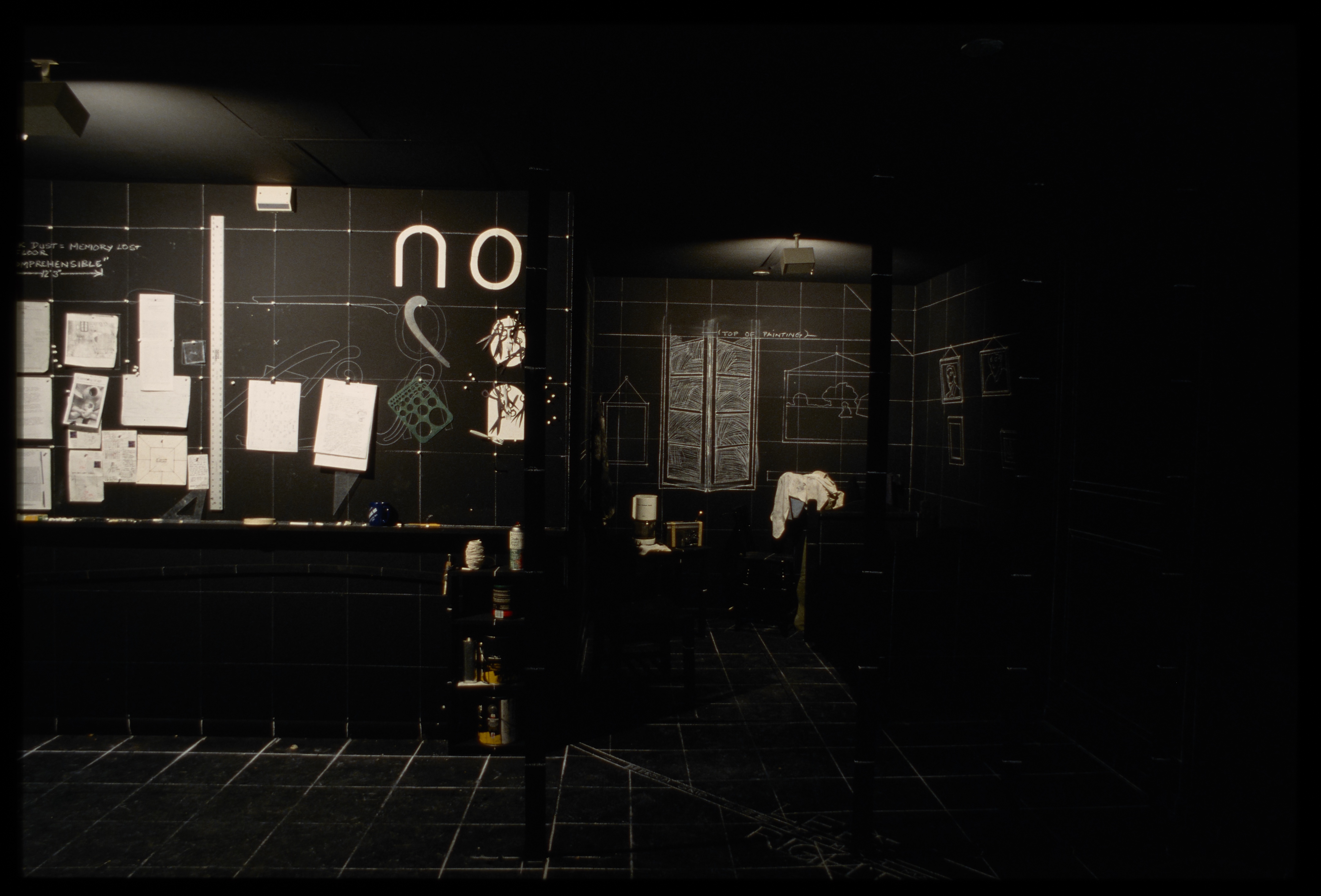 Black room with chalk drawings of furniture to the right, and diagrams and papers hung on the wall to the left. White chalk grid lines entire space.