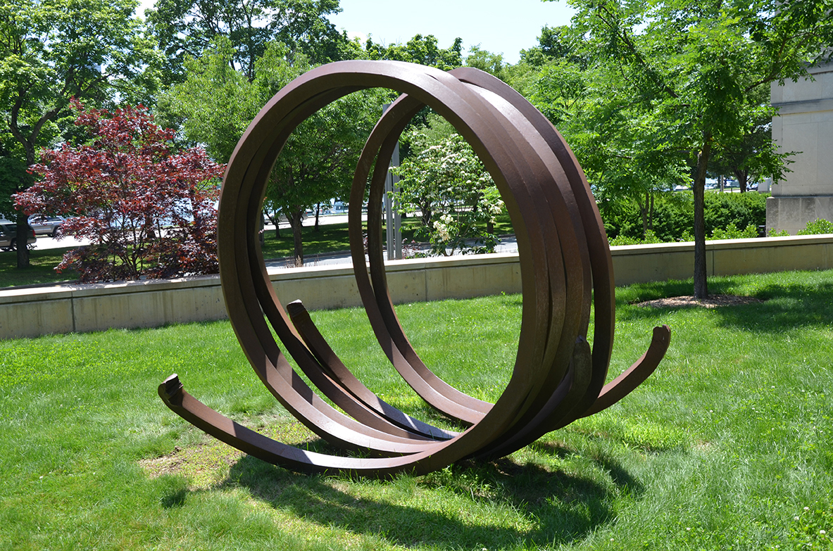 Resting on a sunny lawn, two bands of coiled industrial steel, spiral into a large circular form, resembling a metal “spring