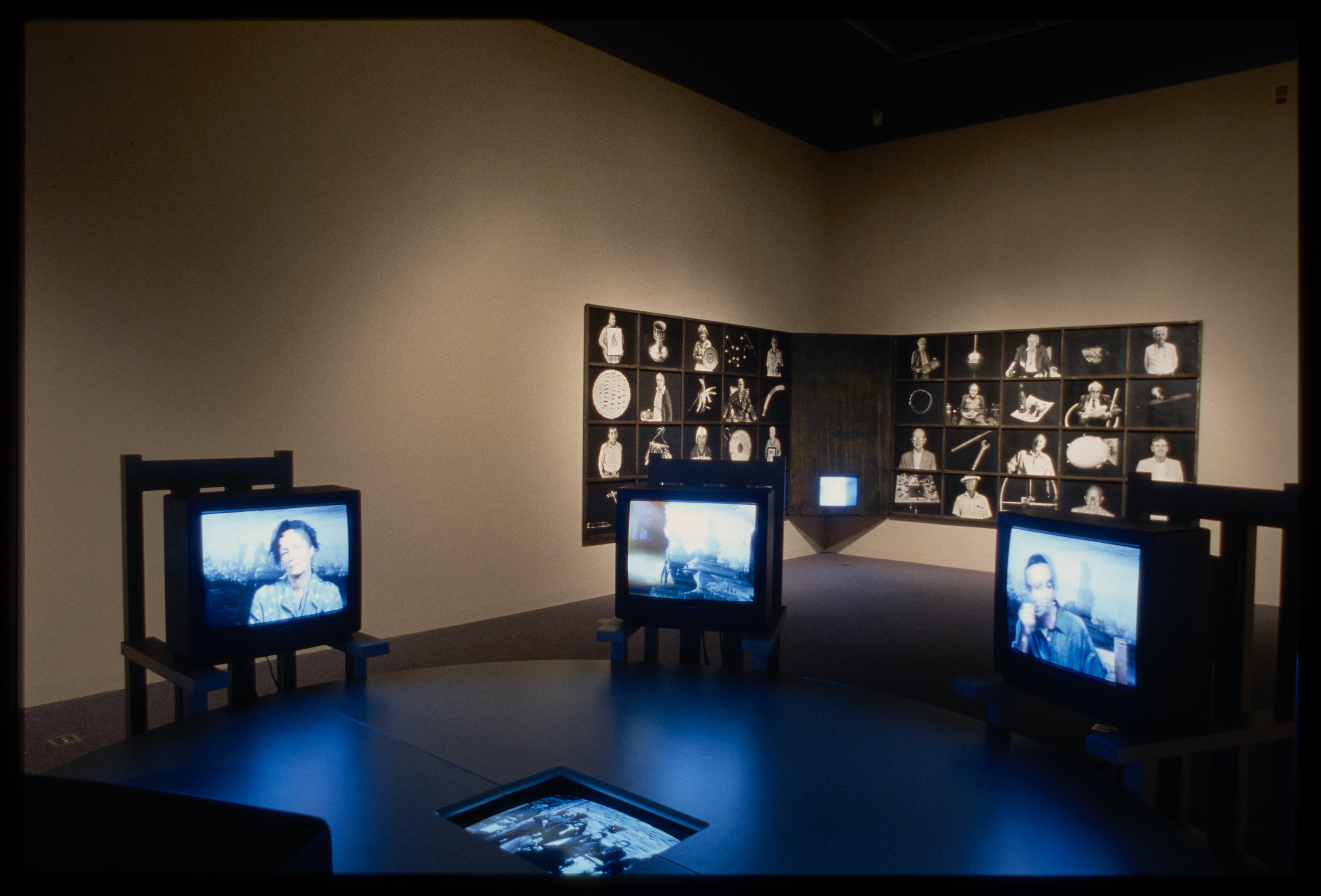 Three tvs with videos of people sit in ring in front of black and white photos of people and items covering the wall corner.