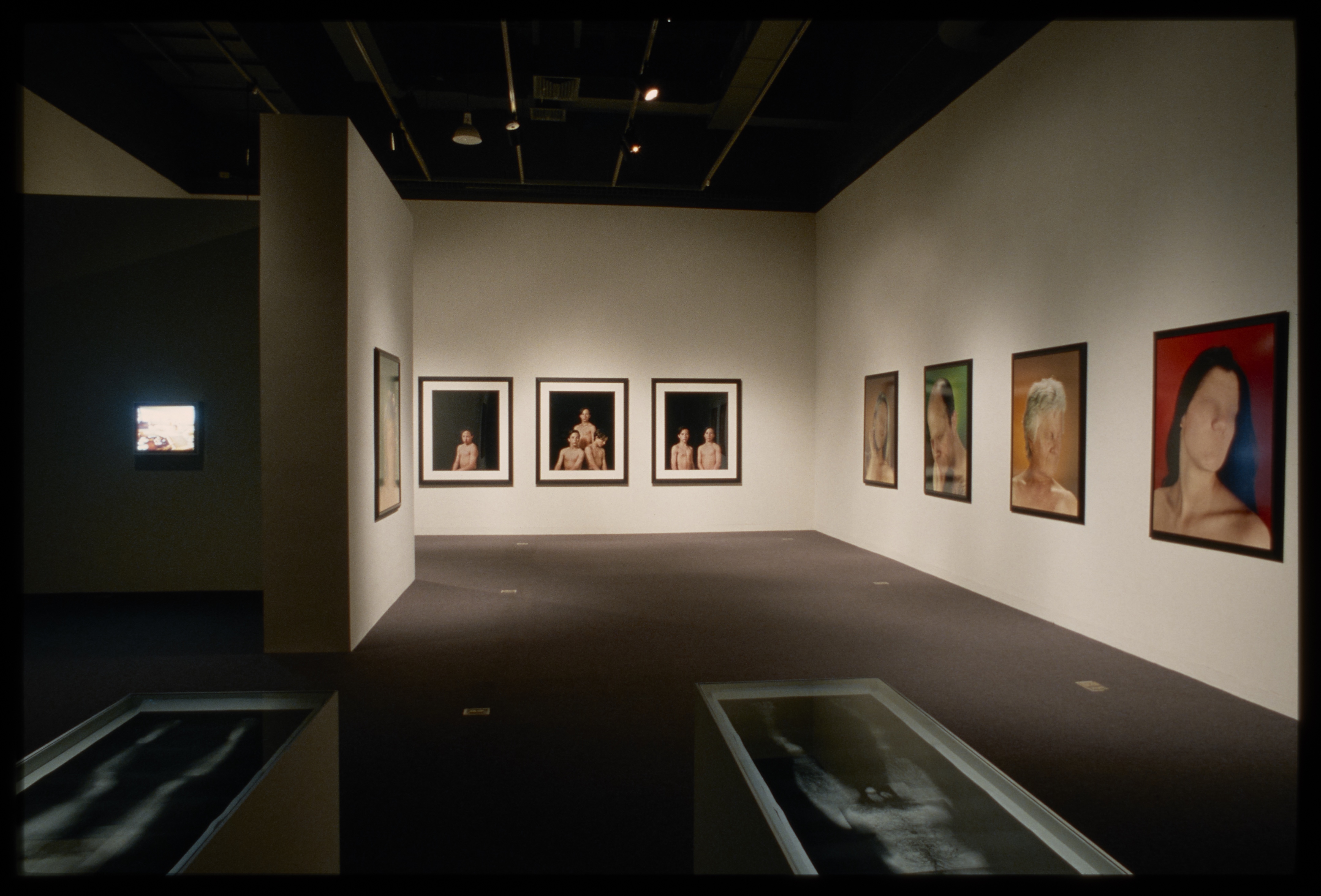 Black and white nude images sit in front of one lit screen and eight headshots of people without eyes or mouths hung on walls.