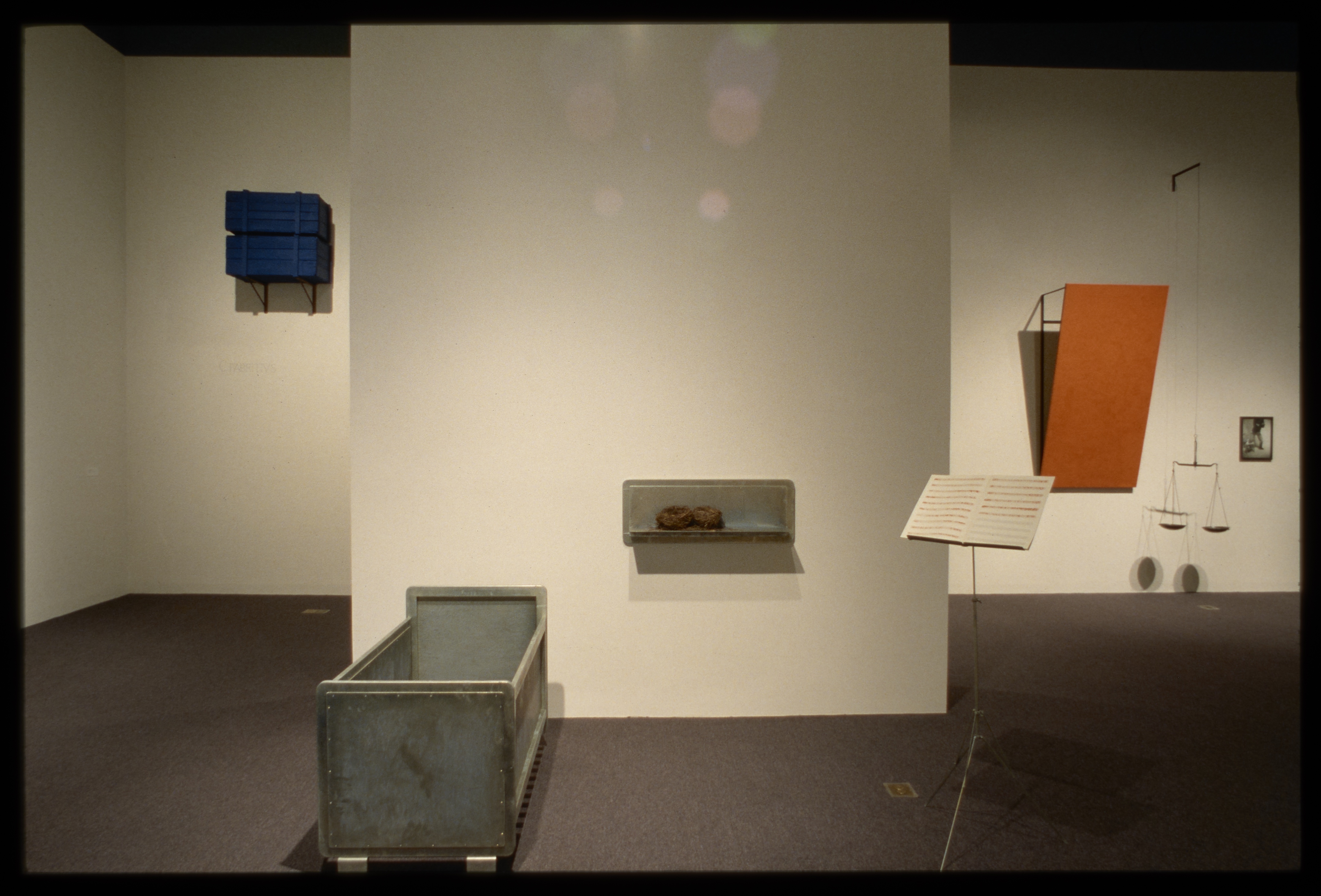 Blue wooden crates hanging on left wall, metal crib, shelf, and music stand in middle, orange work and hanging scales on right.