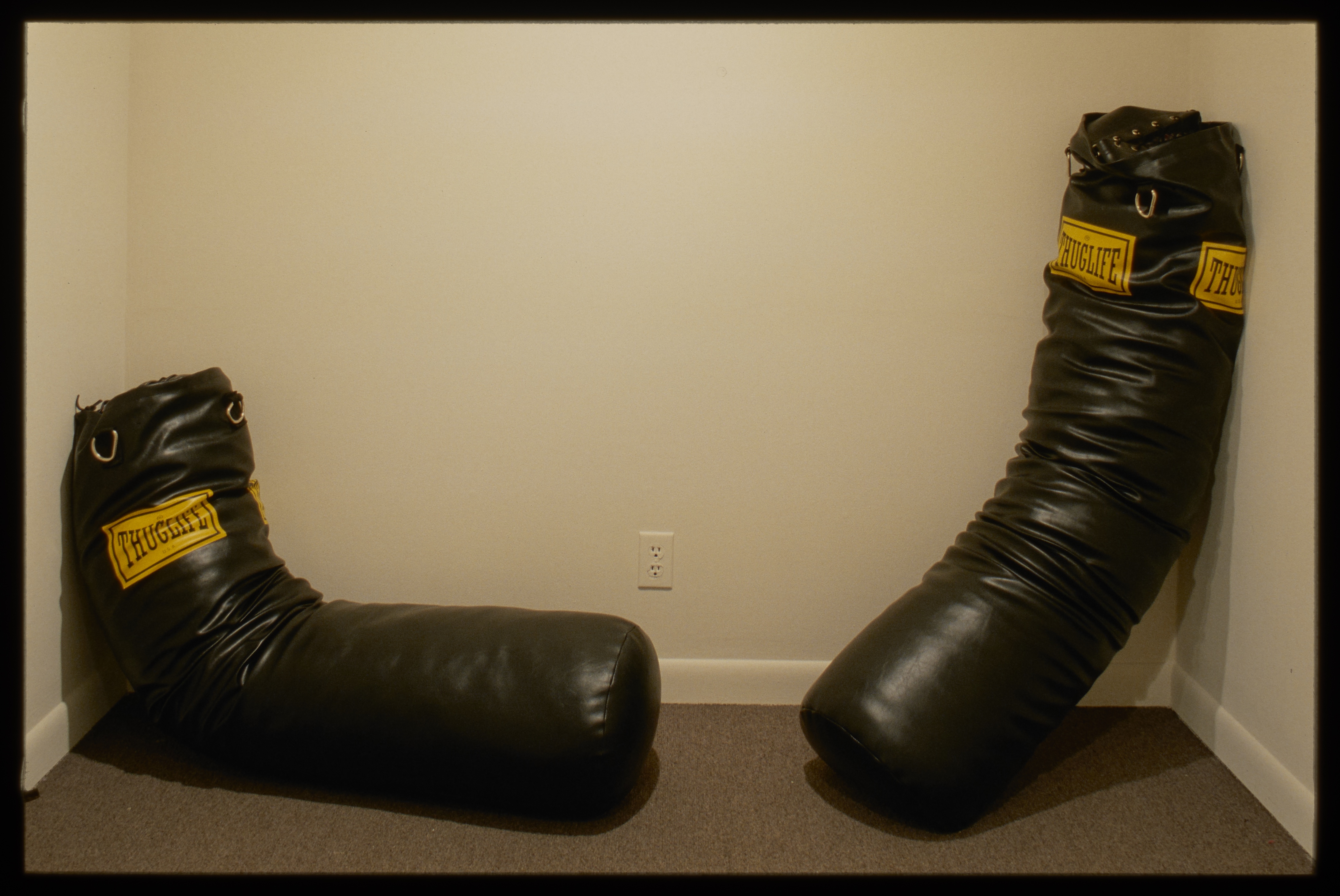 Two black punching bags with yellow logo reading “Thuglife” sit slumped across from eachother on gallery wall