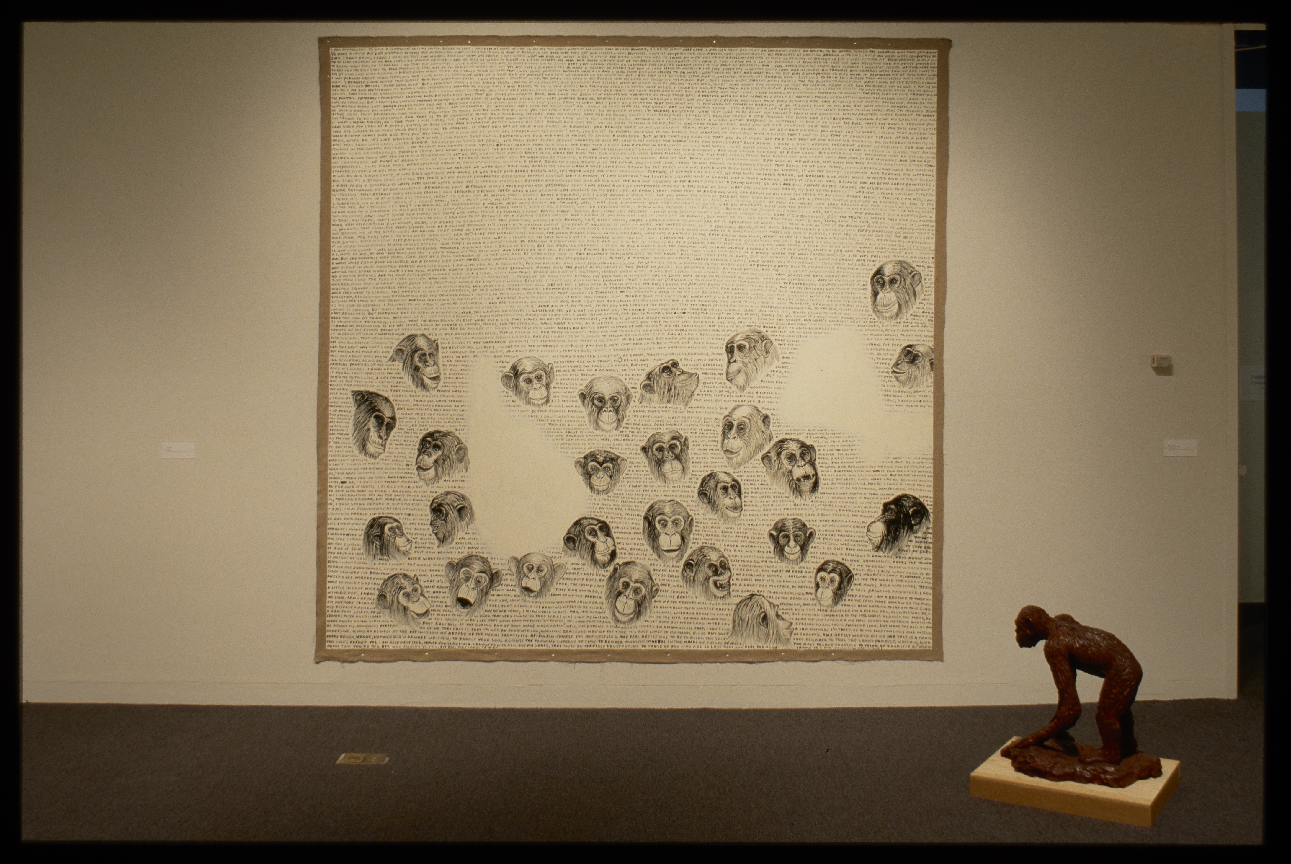 Brown clay ape sculpture stands in front of large wall mural with inked apes heads with various facial expressions and text.