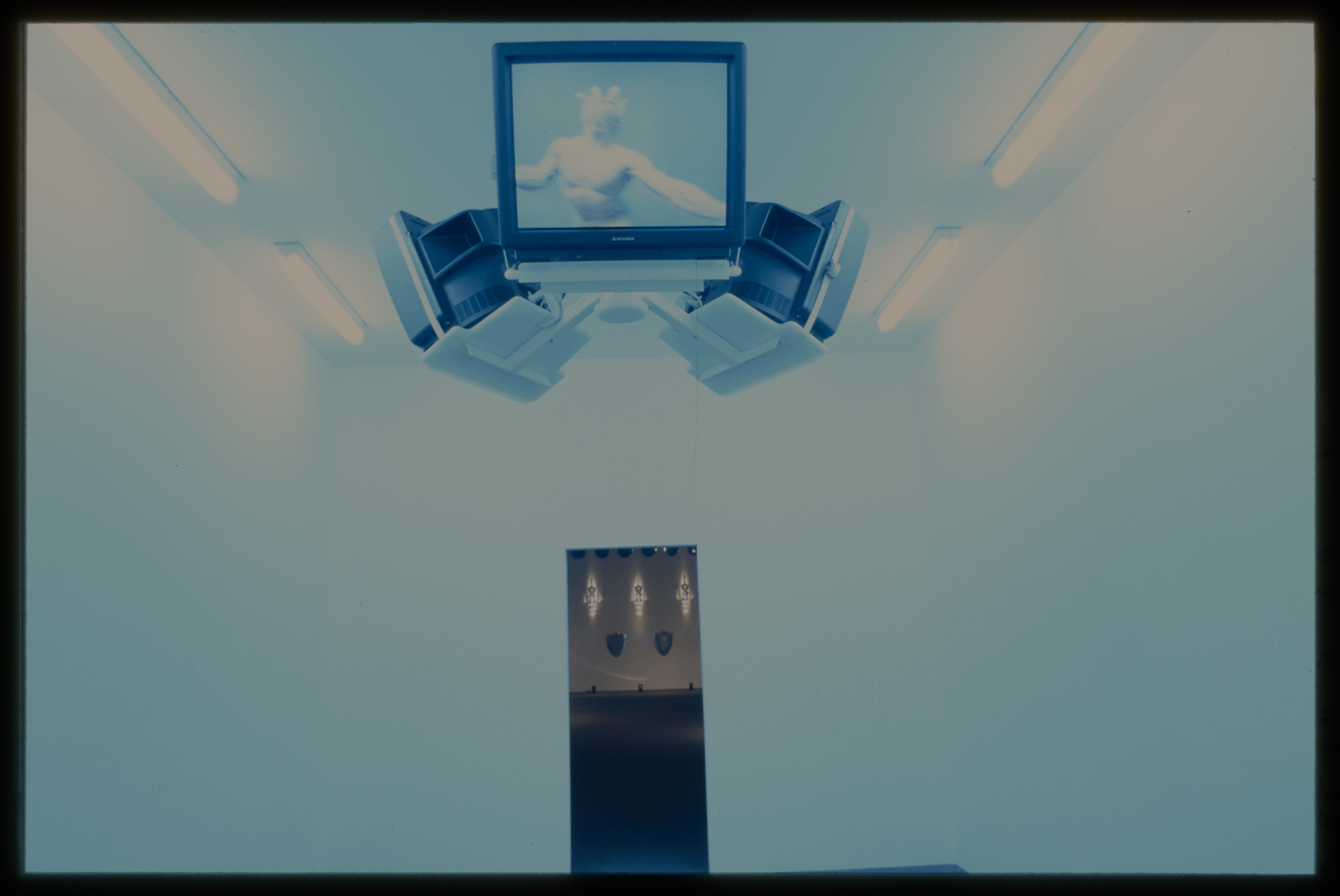 A room with three monitors hanging from the ceiling. Fluorescent lighting gives the image a blurry filter of bright blue.