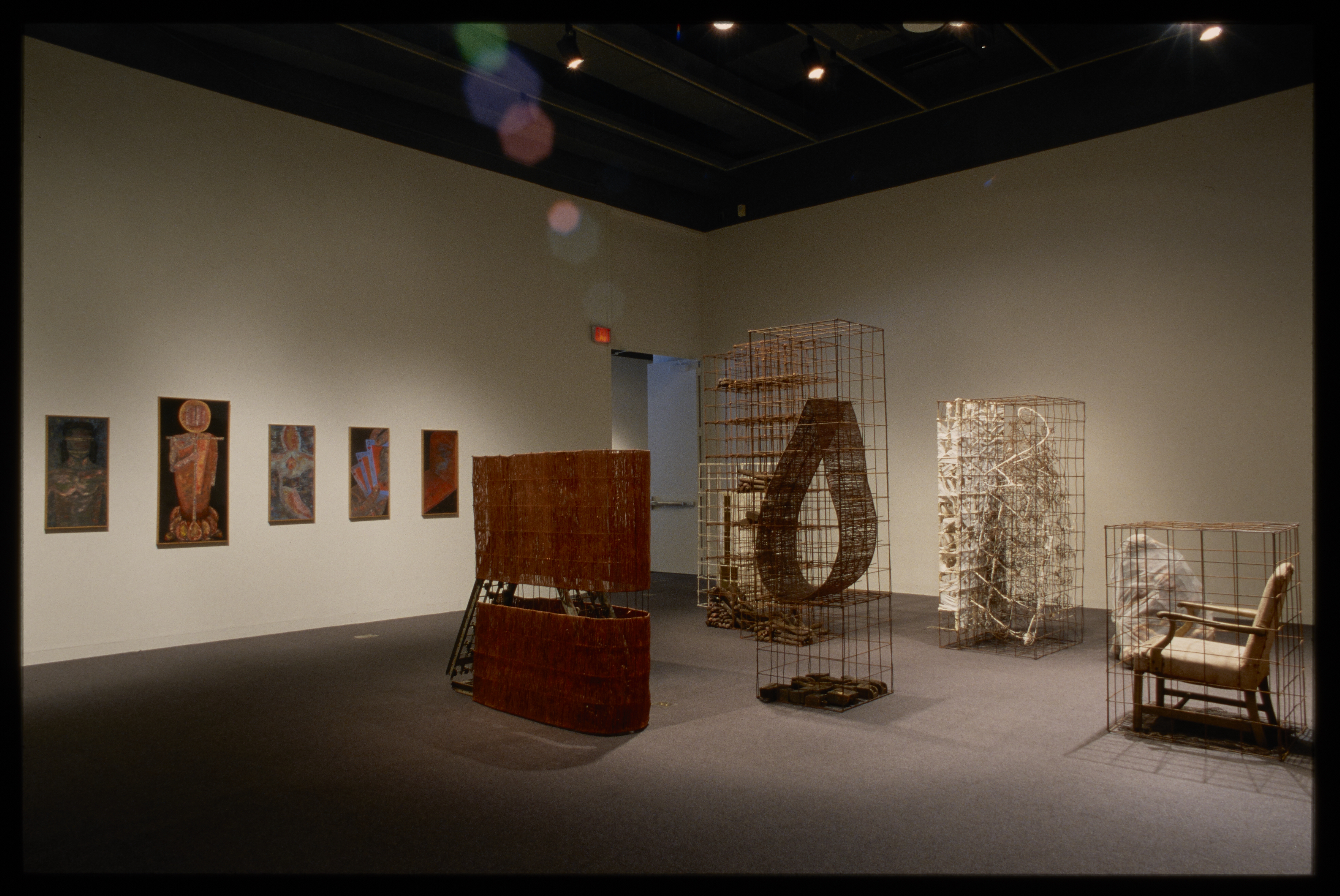 Various sculptures and objects are caged in the center of the gallery. Figurative paintings overlook from the opposing wall.