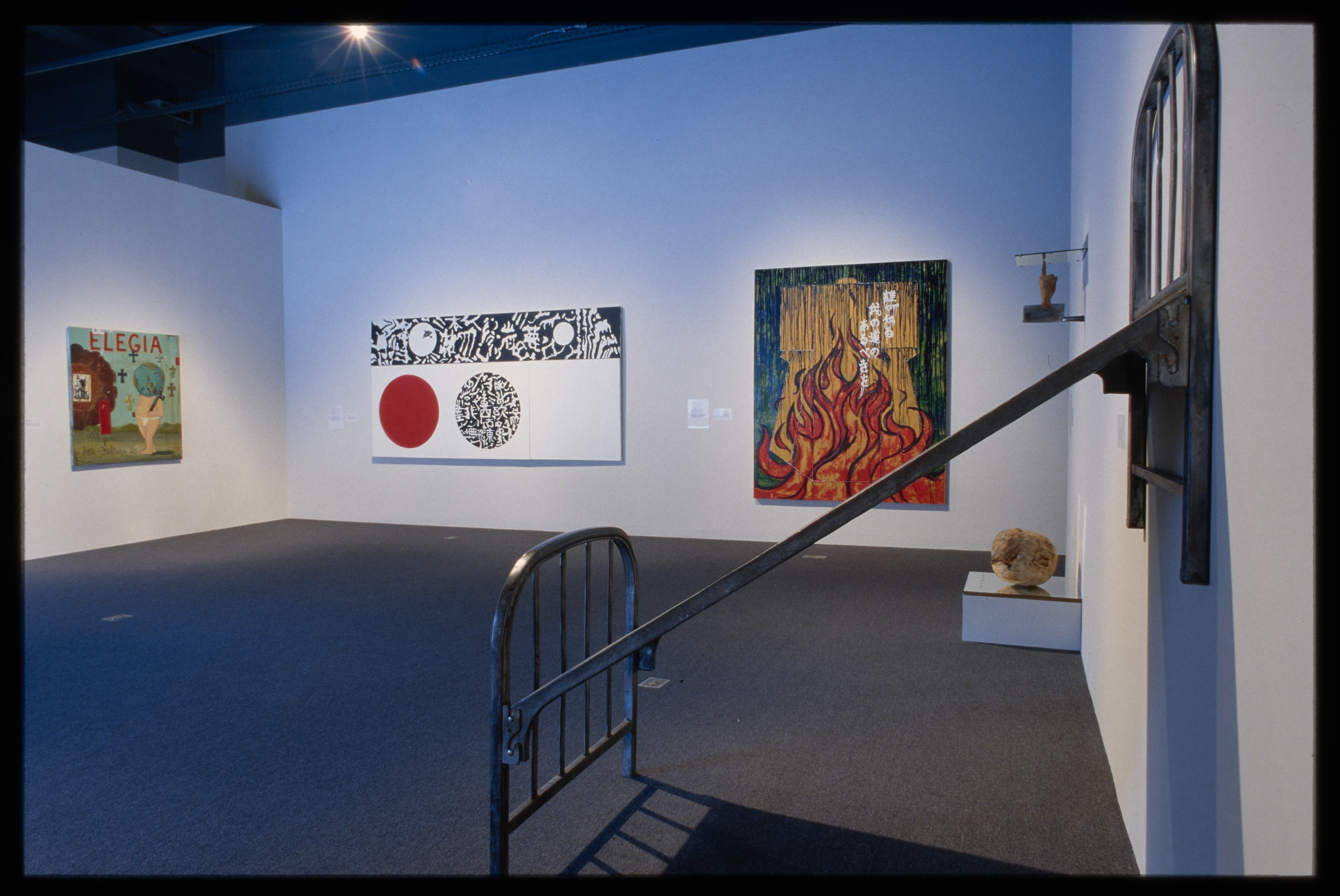 A metal bed frame is hoisted onto the wall. Its headboard is angled diagonally from the base with other works just behind. 