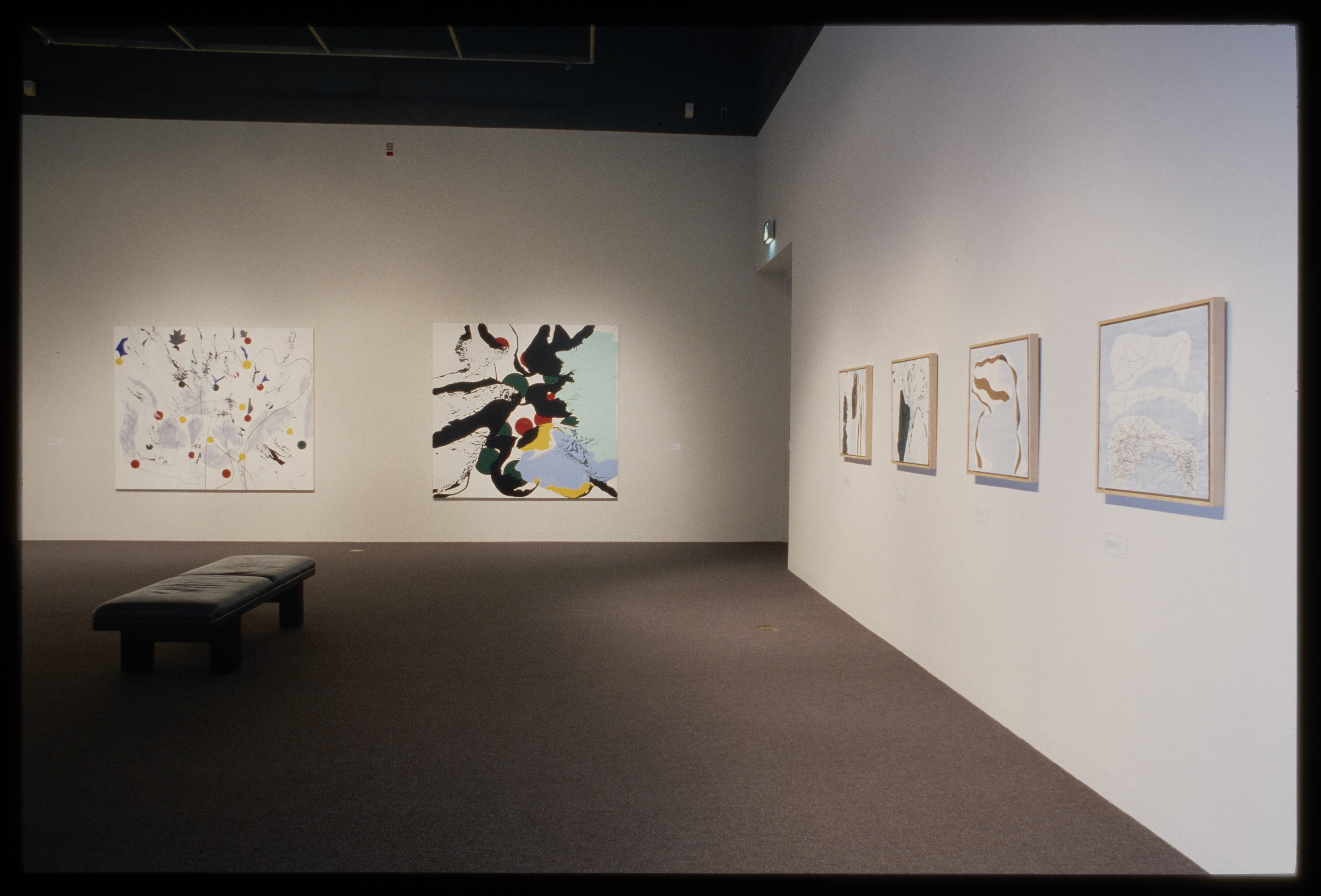 Two large and four small abstract paintings with white background and subtle colorful splotches and dots hang on adjacent walls.