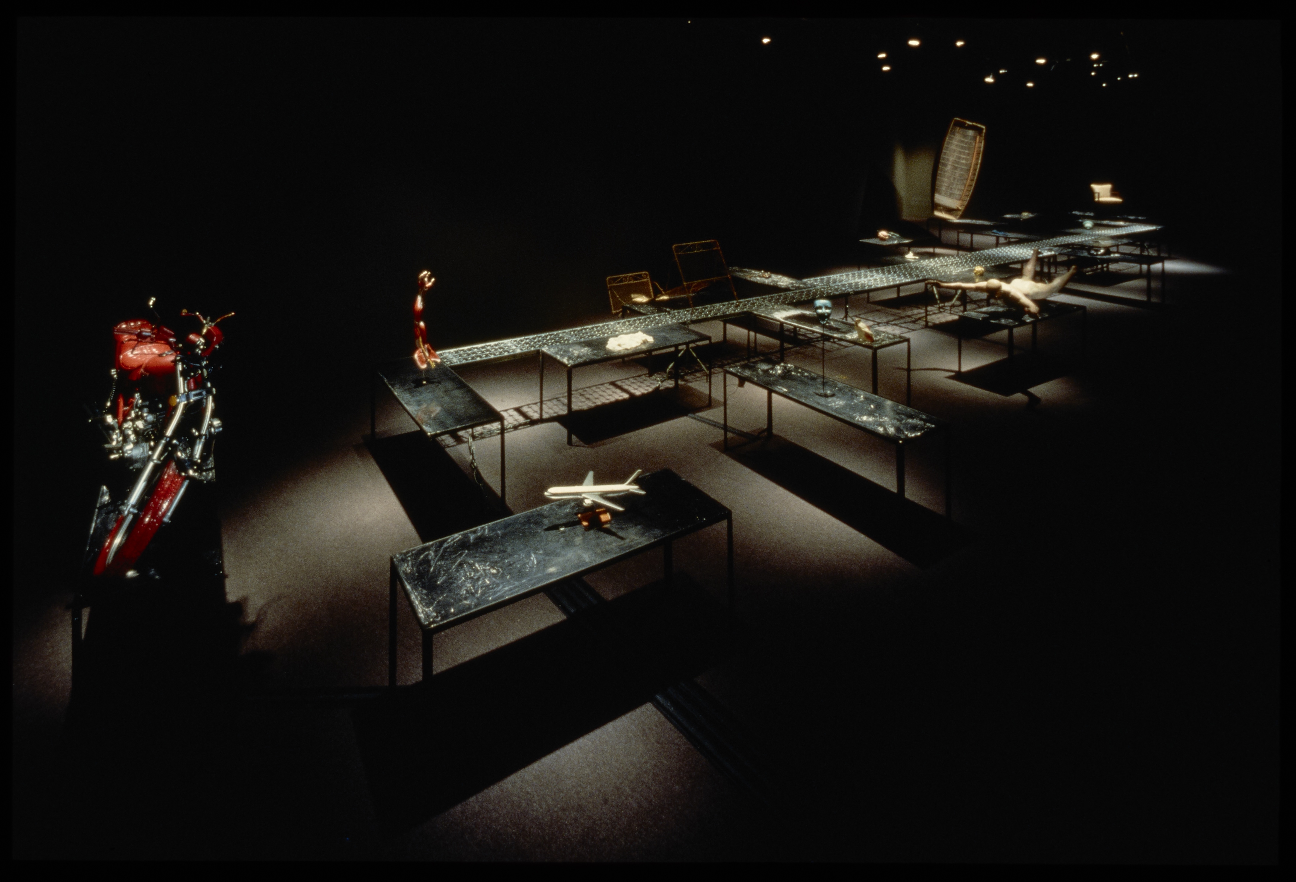 Dark gallery with spotlit motorcycle, chairs, and maze of tables with found objects on them.