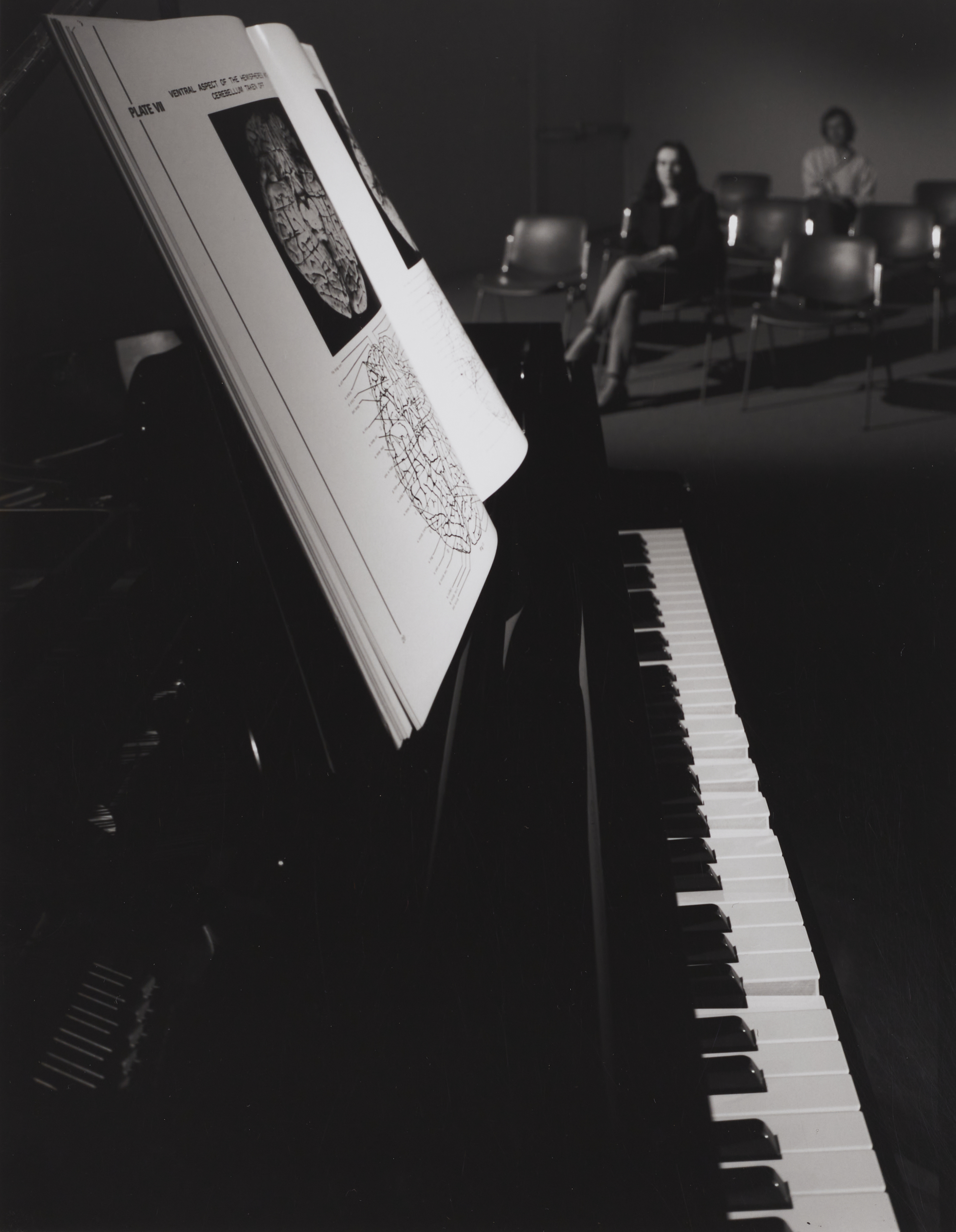 Keyboard of black grand piano with book of brain images in foreground, 2 blurred people sitting in chairs in background