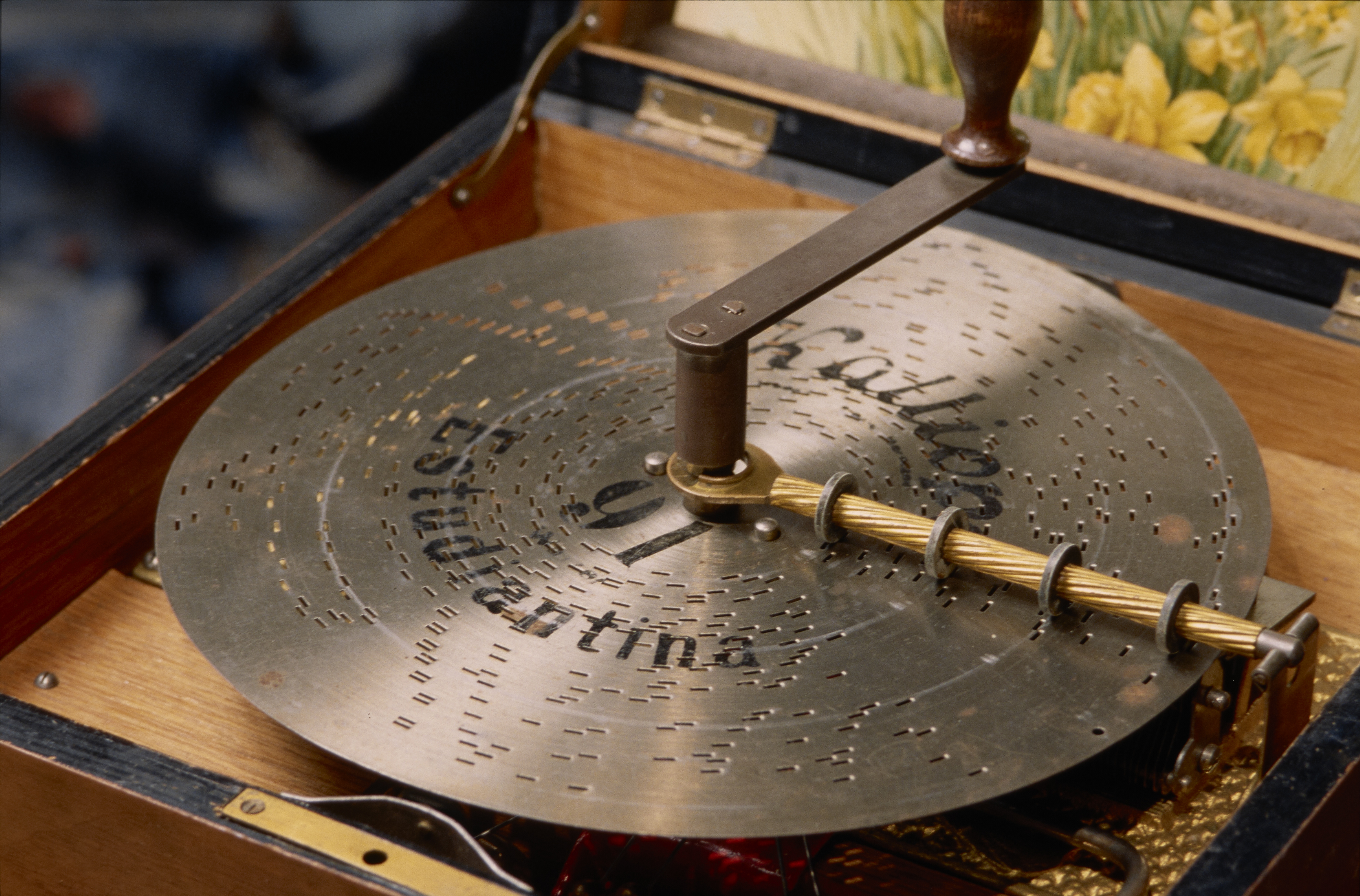 Steel disc punched with rectangular holes, beneath a brown-metal and wood crank, fitted in a wood box