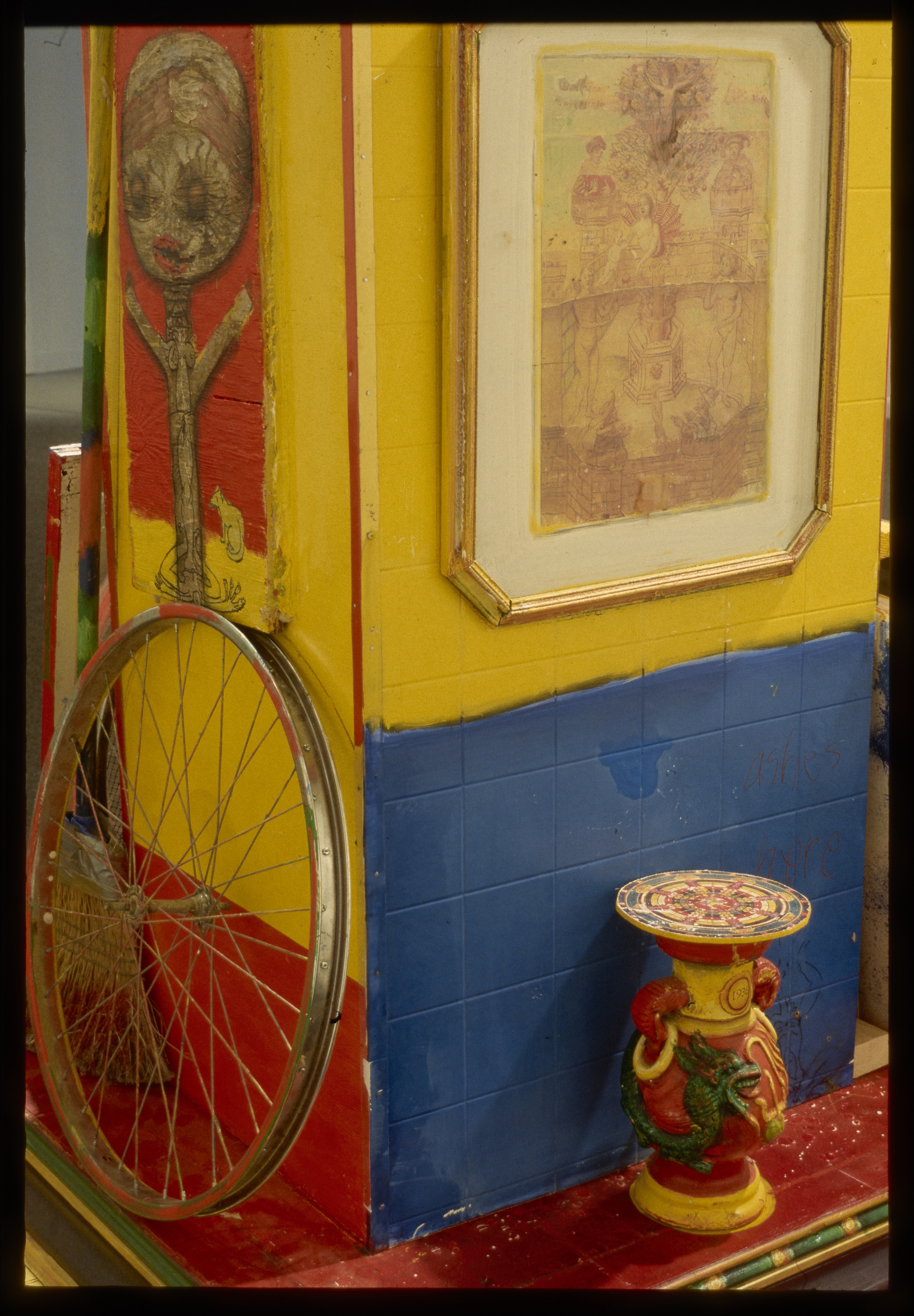 Detail of an image with bright yellow and blue walls with a bicycle wheel and a small table on view.