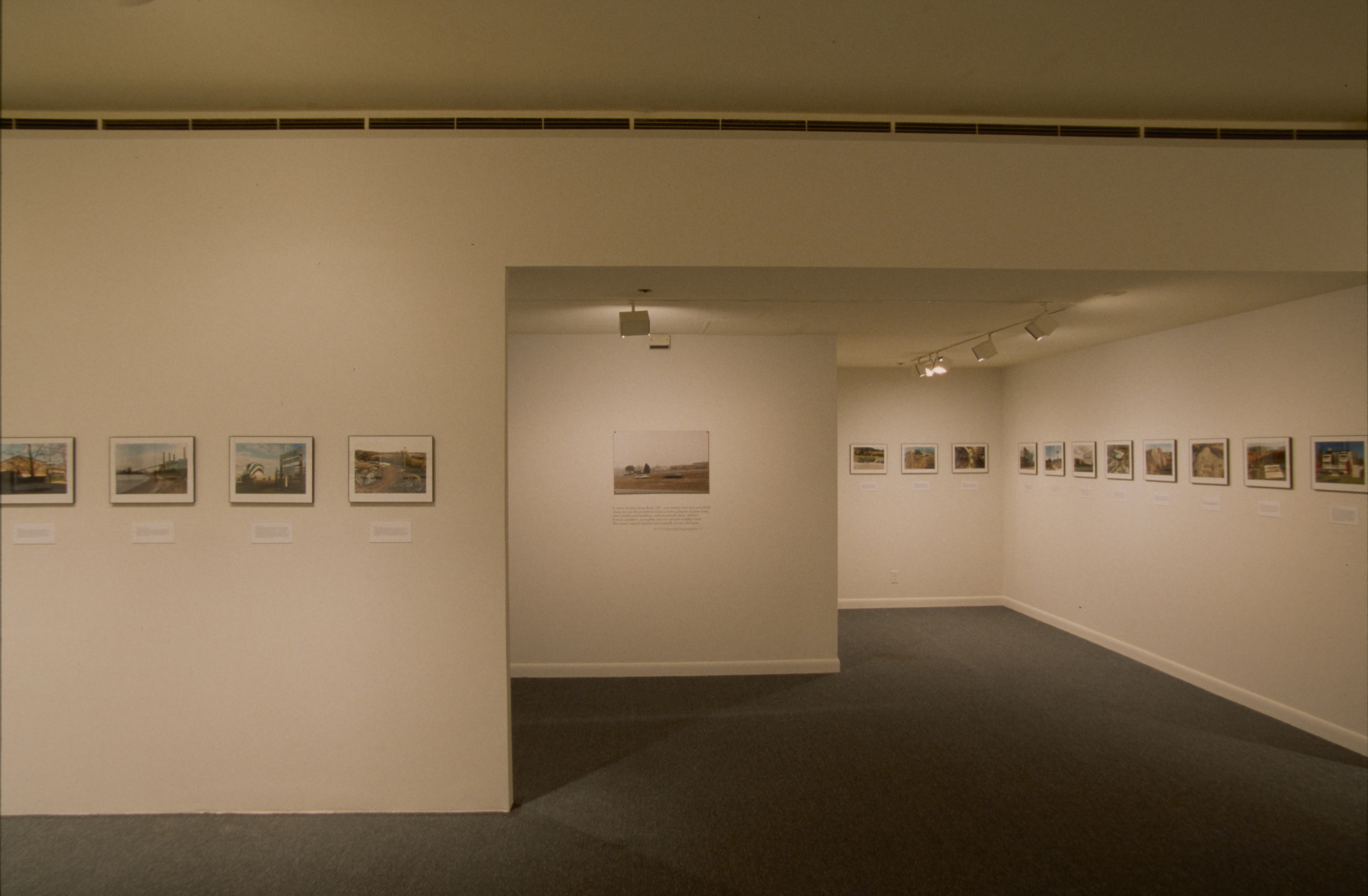 A row of framed color photographs hangs at eye level on white walls around 3 rooms of the gallery.