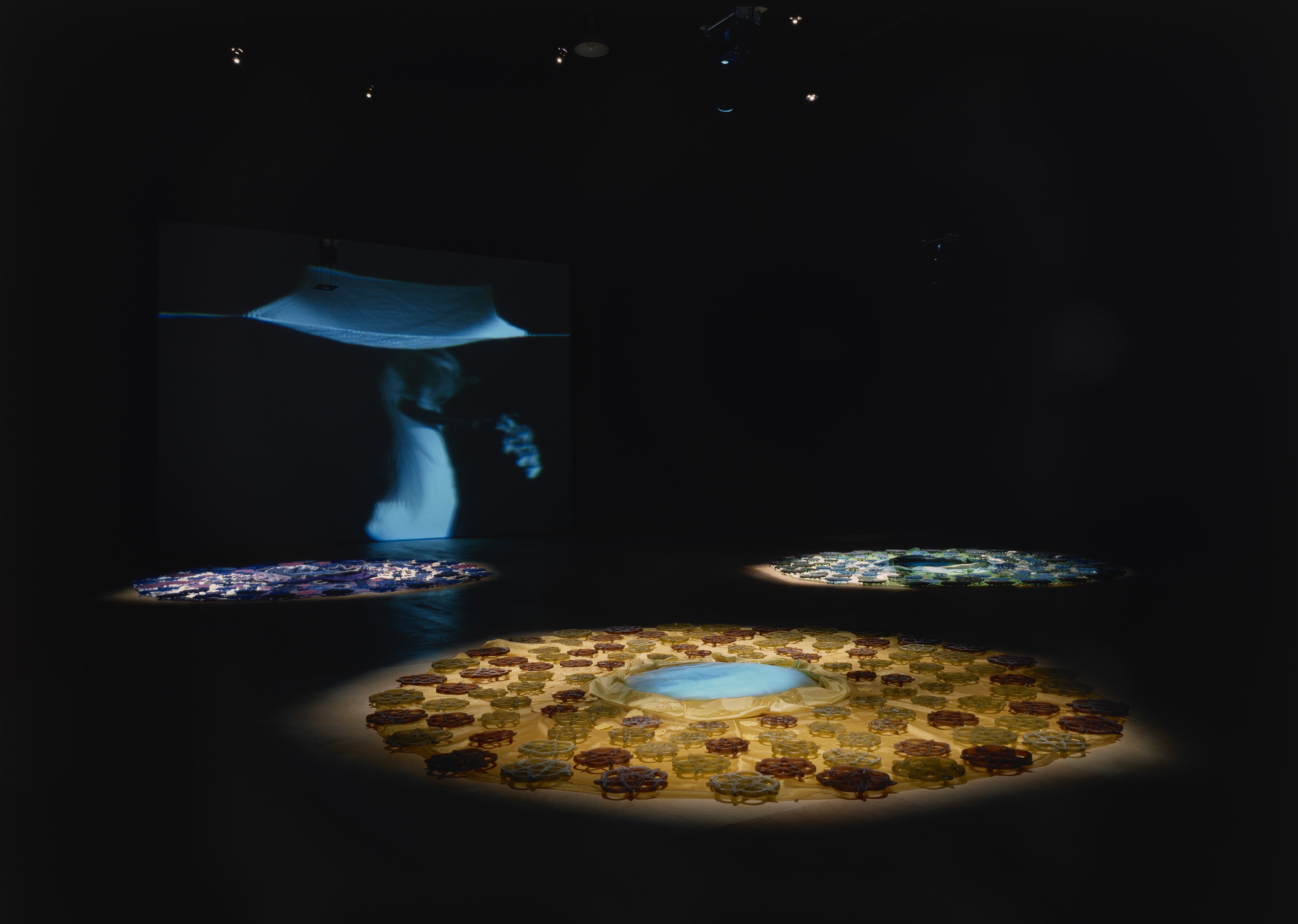Blue, green glass trivets and flowers on shiny tan fabric seem to swirl around an iridescent blue-green disc on a dark floor.
