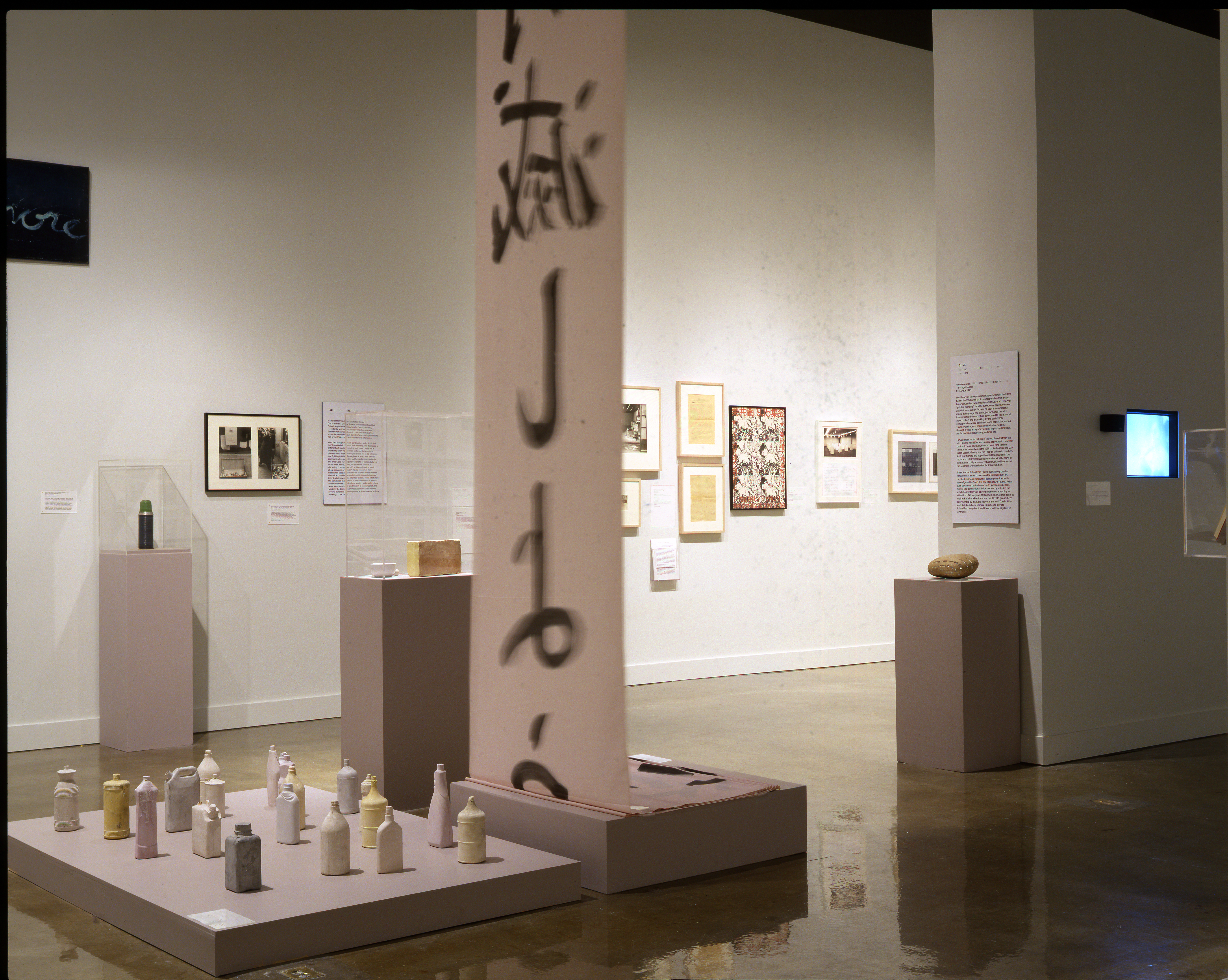 Muted colored bottles on a base, calligraphy on a large hanging scroll, art on walls and pedestals in the space beyond.