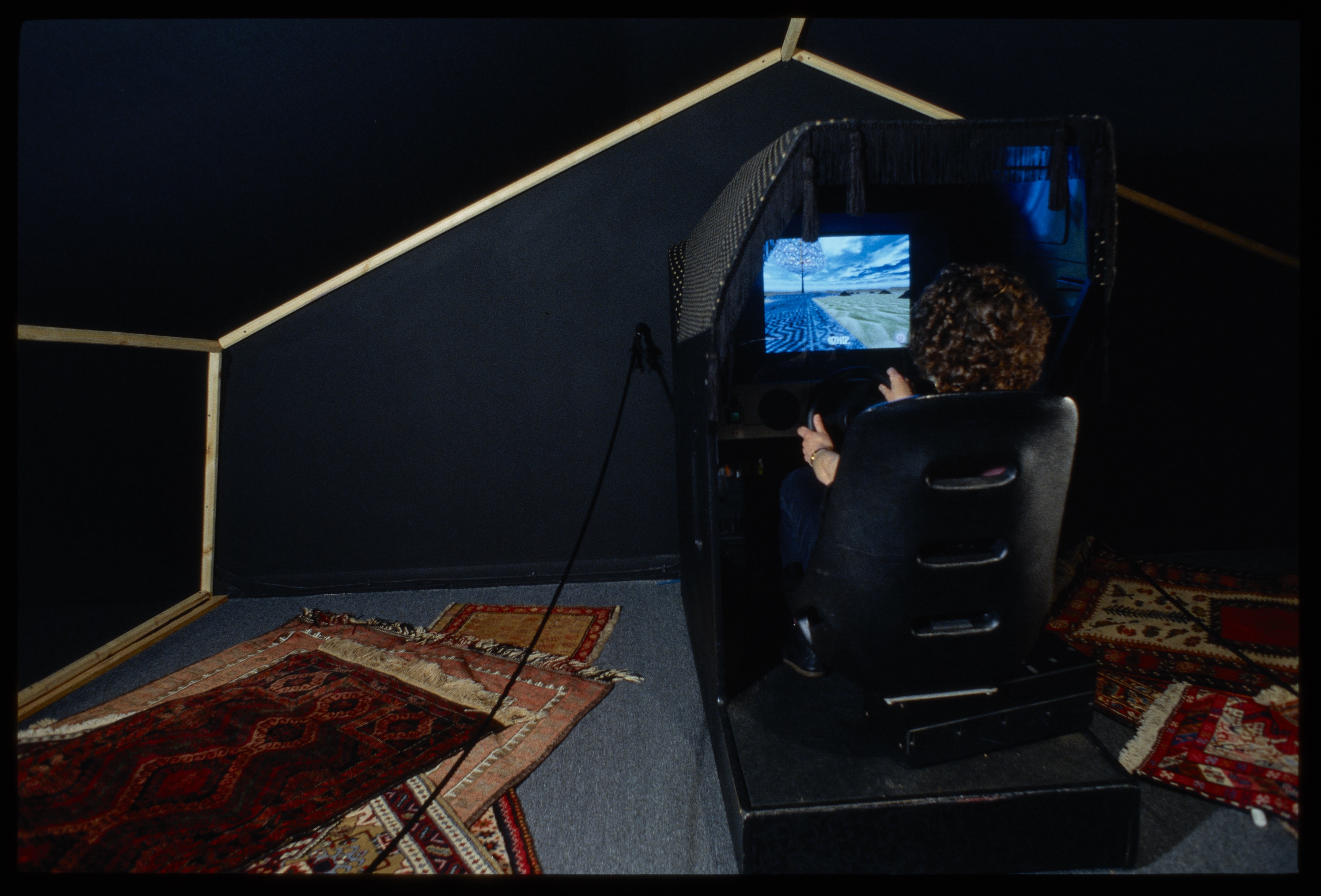 A woman sitting in a chair that resembles an arcade game with a computer monitor in front.