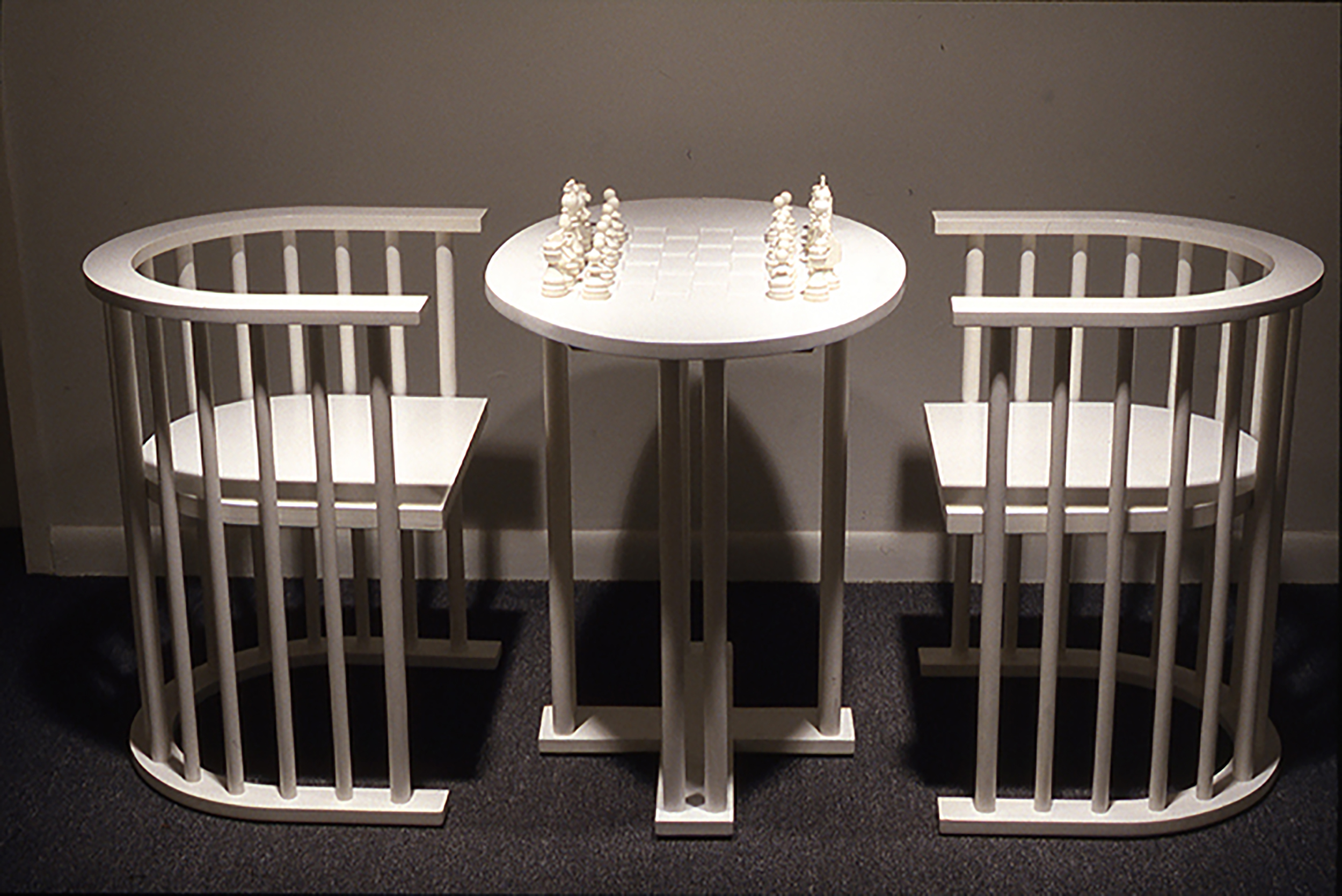 Straight on view of a wooden chess table with pieces set up and 2 wooden chairs, all painted white