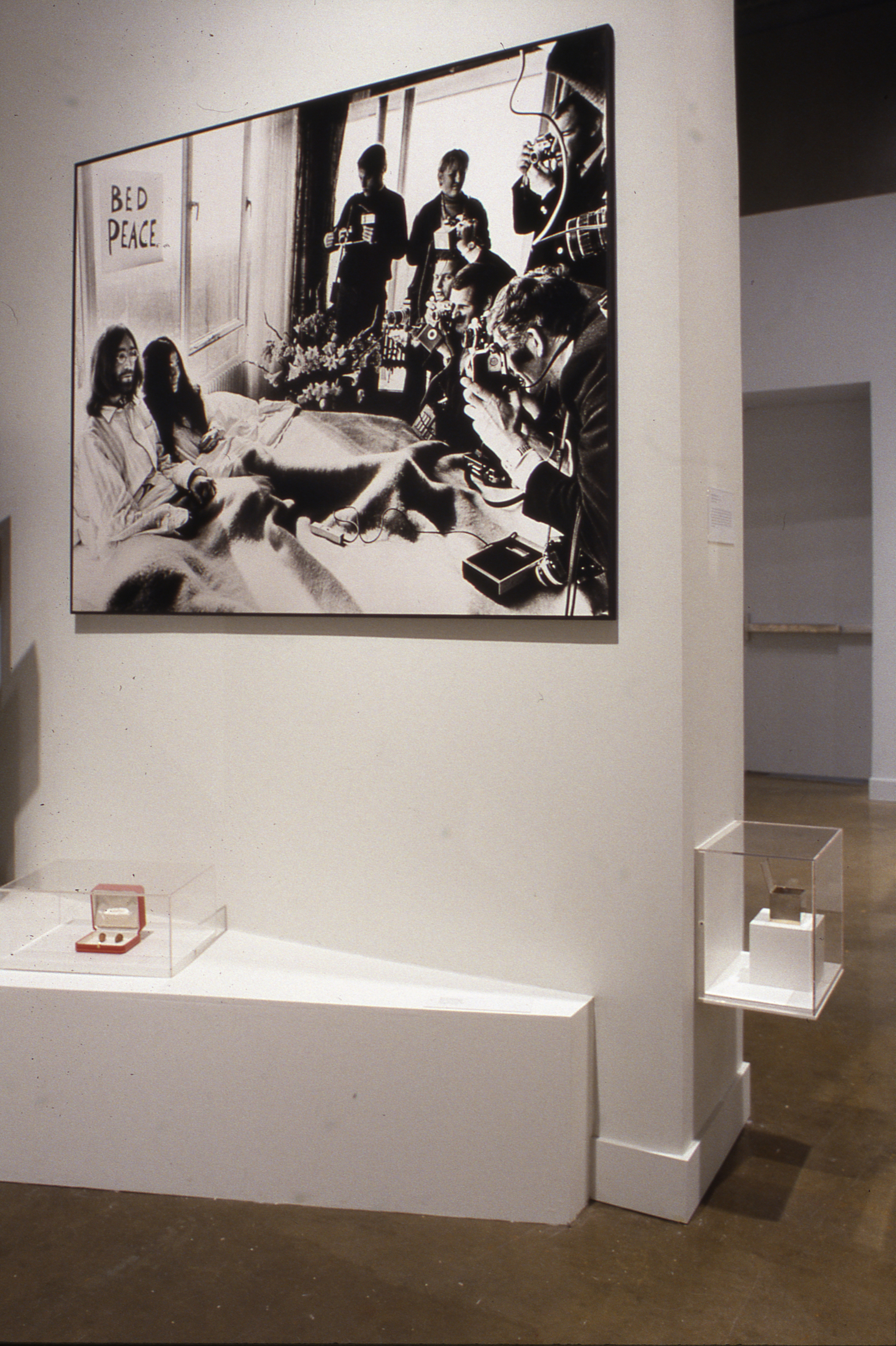 Small vitrines hold objects below enlarged black and white photo of John Lennon and Yoko Ono in bed surrounded by the media